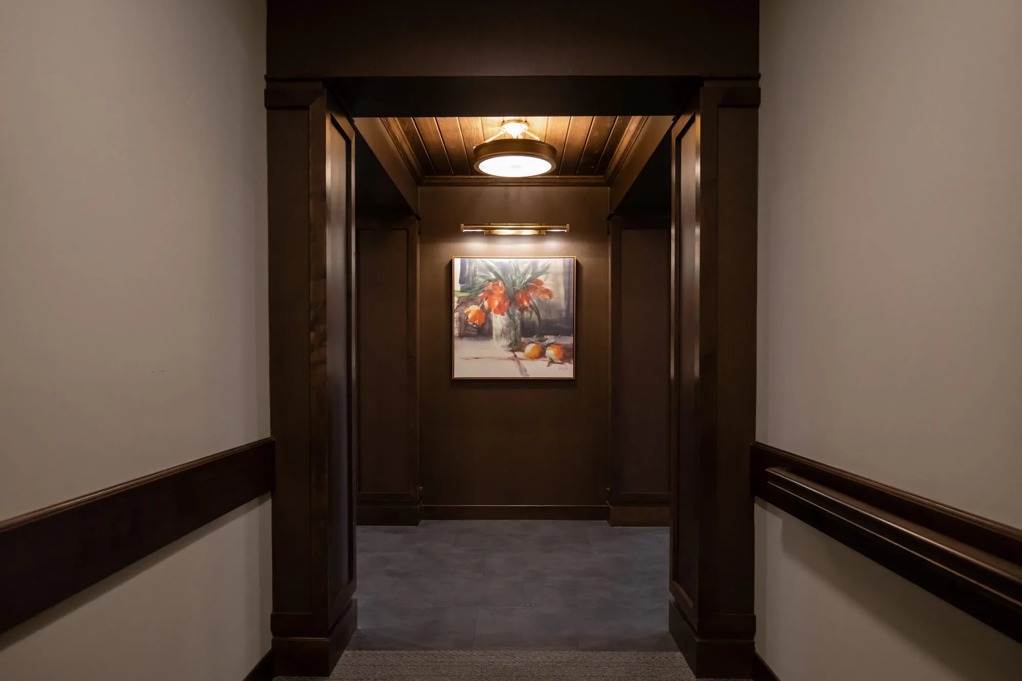 Active adult community corridor panelled in dark wood, opening to an alcove with a still life painting