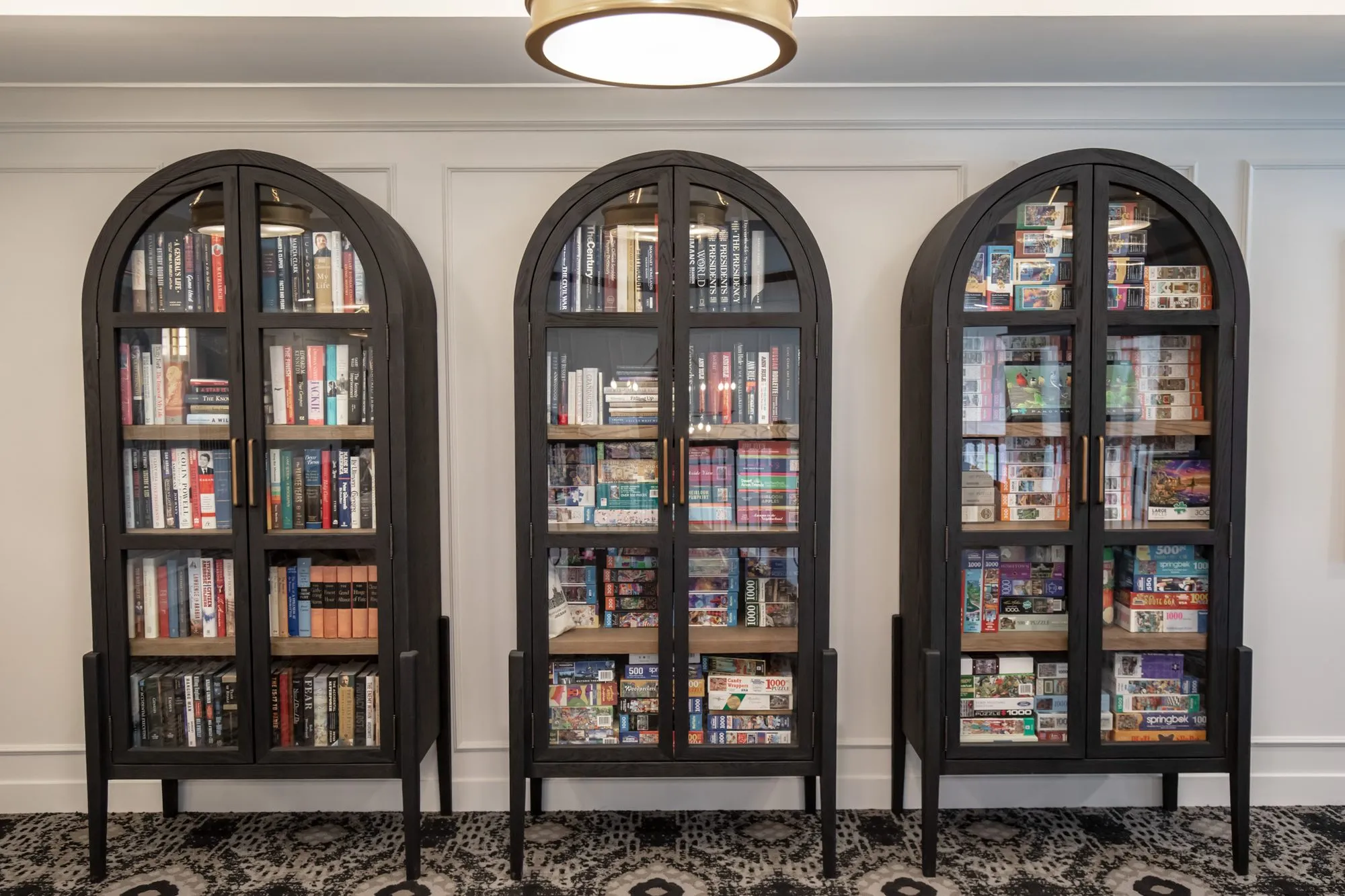 Active adult community interior detail: three black arched glass cabinets filled with books against a white panelled wall