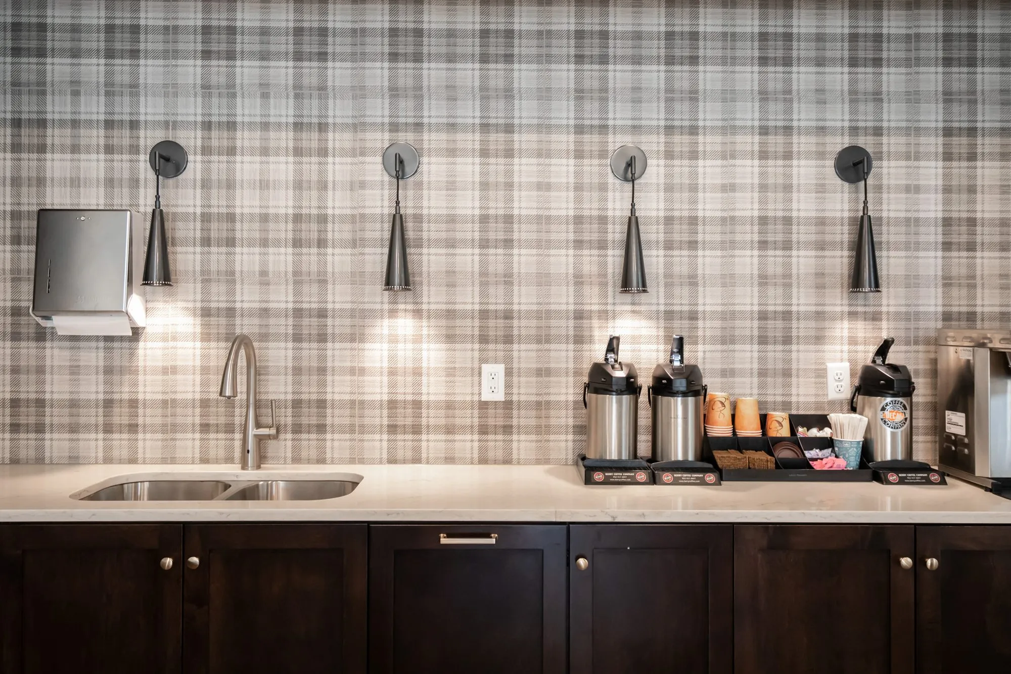 Active adult community coffee bar with dark cabinetry, black cone sconces and beverage dispensers against plaid wallpaper