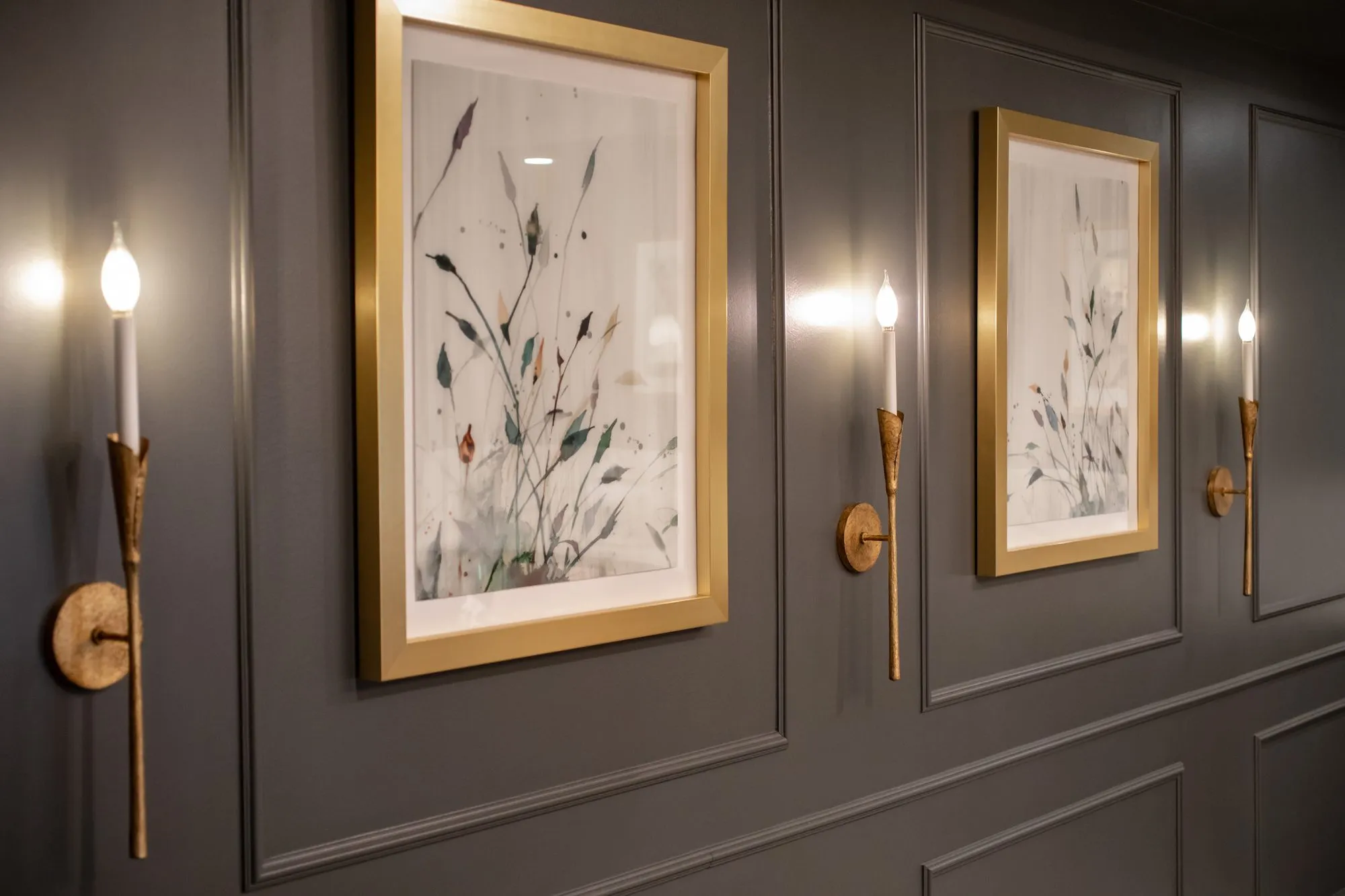 Active adult community interior detail: gold framed botanical prints and gold torch sconces along a grey panelled wall