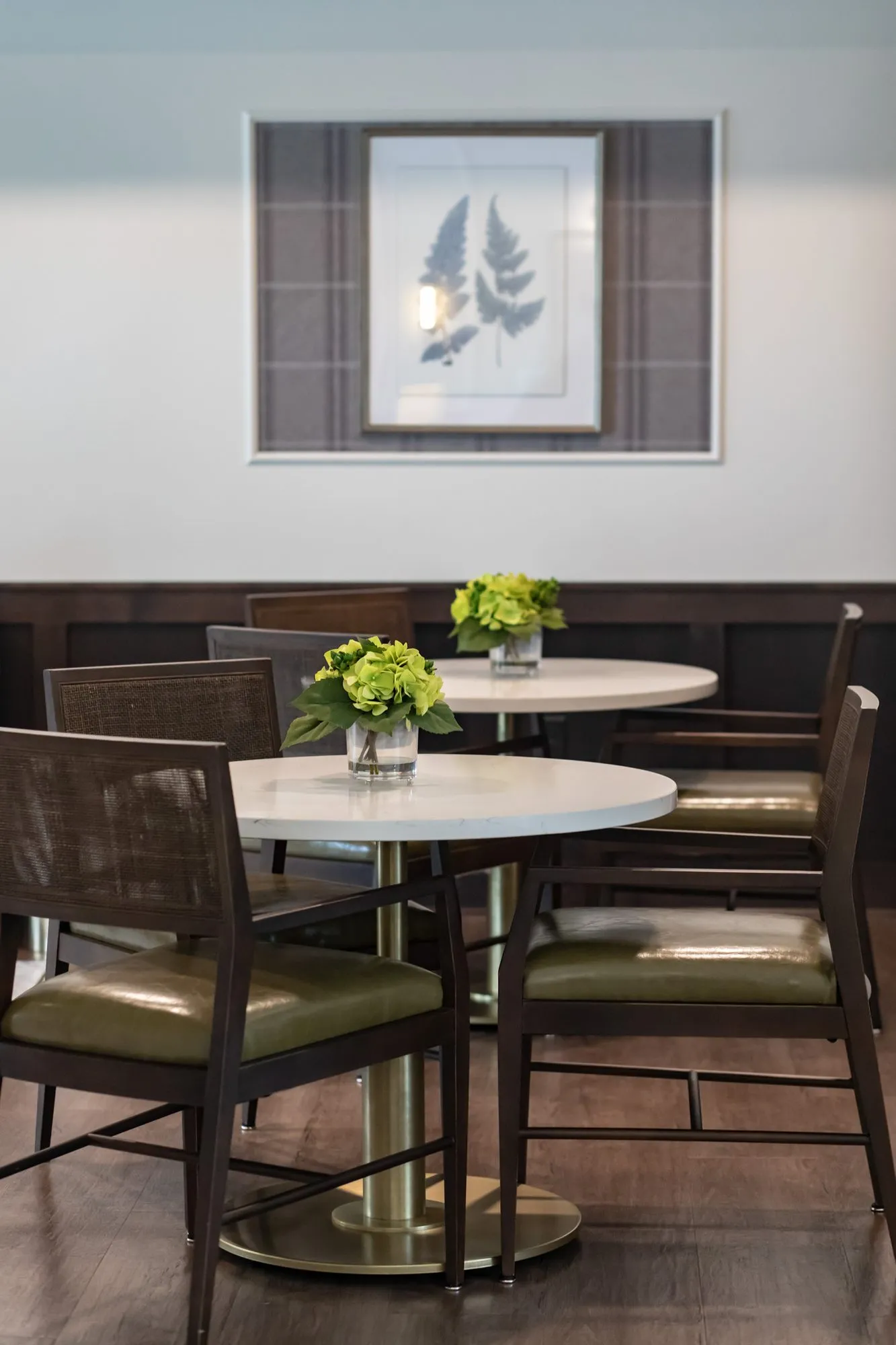 Active adult community interior detail: round cafe tables with cane back chairs and green seats below a fern print