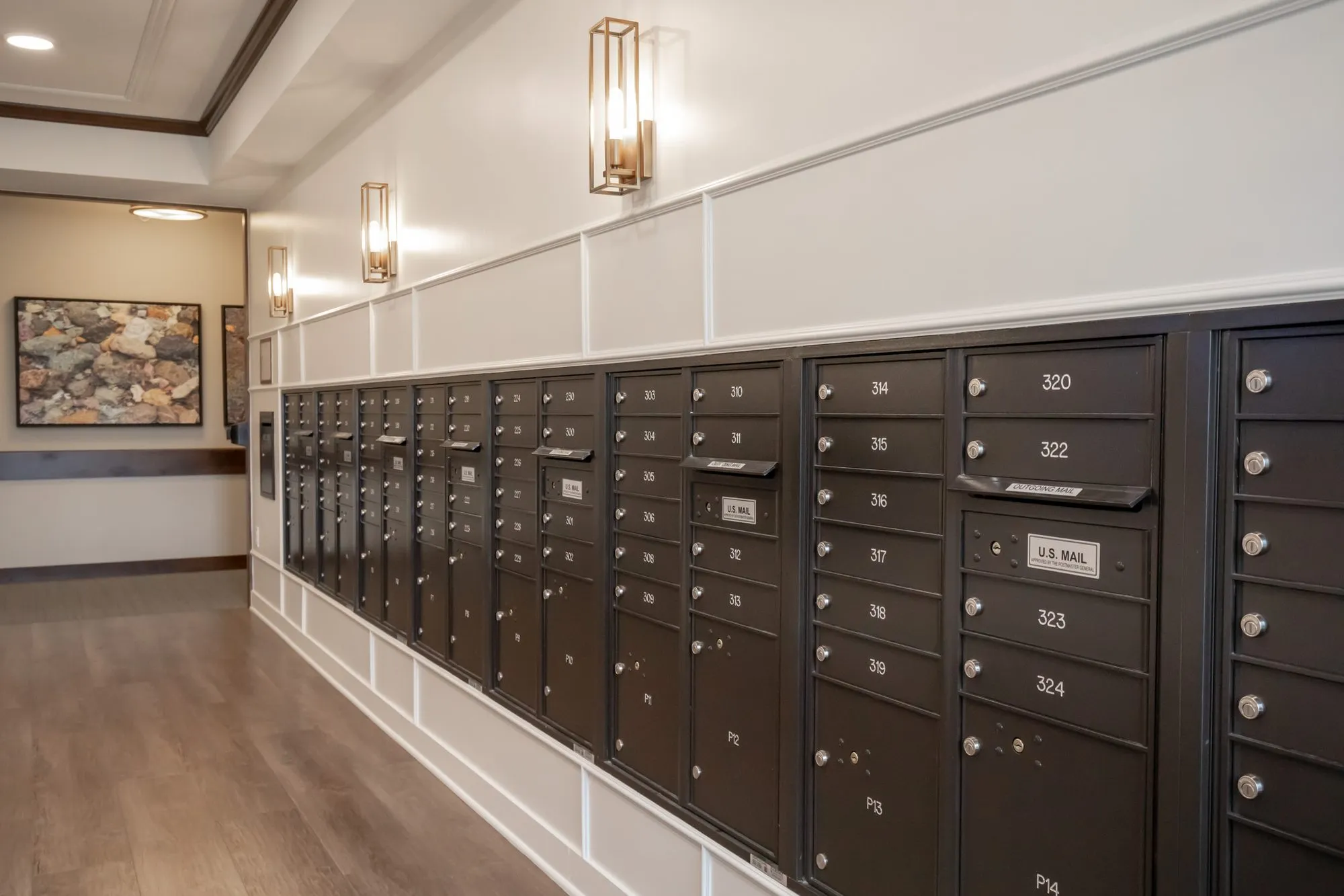 Active adult community mail room with a long run of dark bronze mailboxes below white panelling and lantern sconces