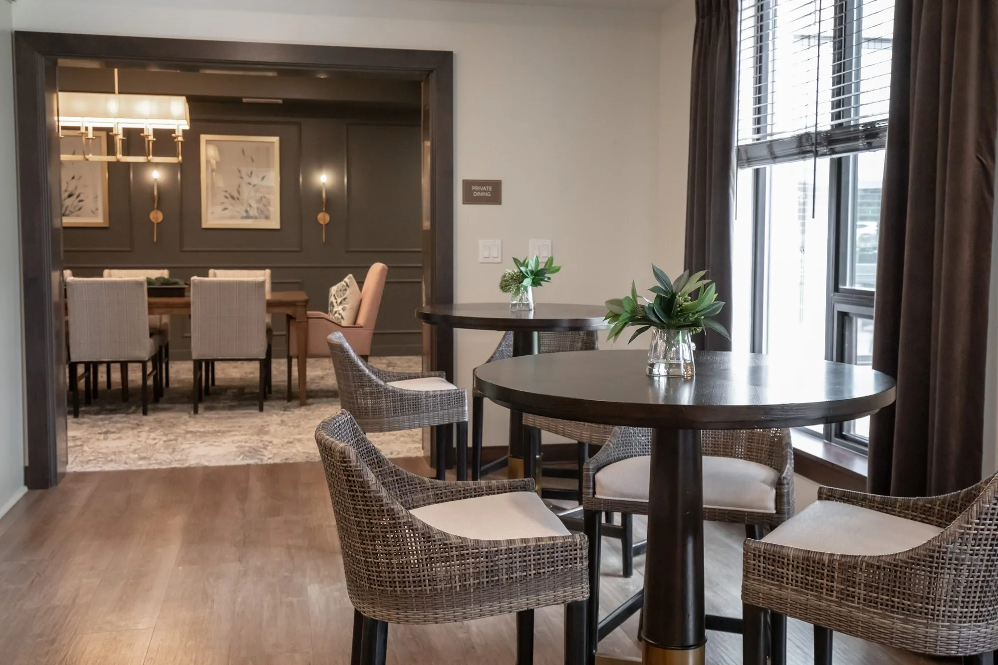 Active adult community dining area with woven chairs around high tables by a window, the private dining room beyond