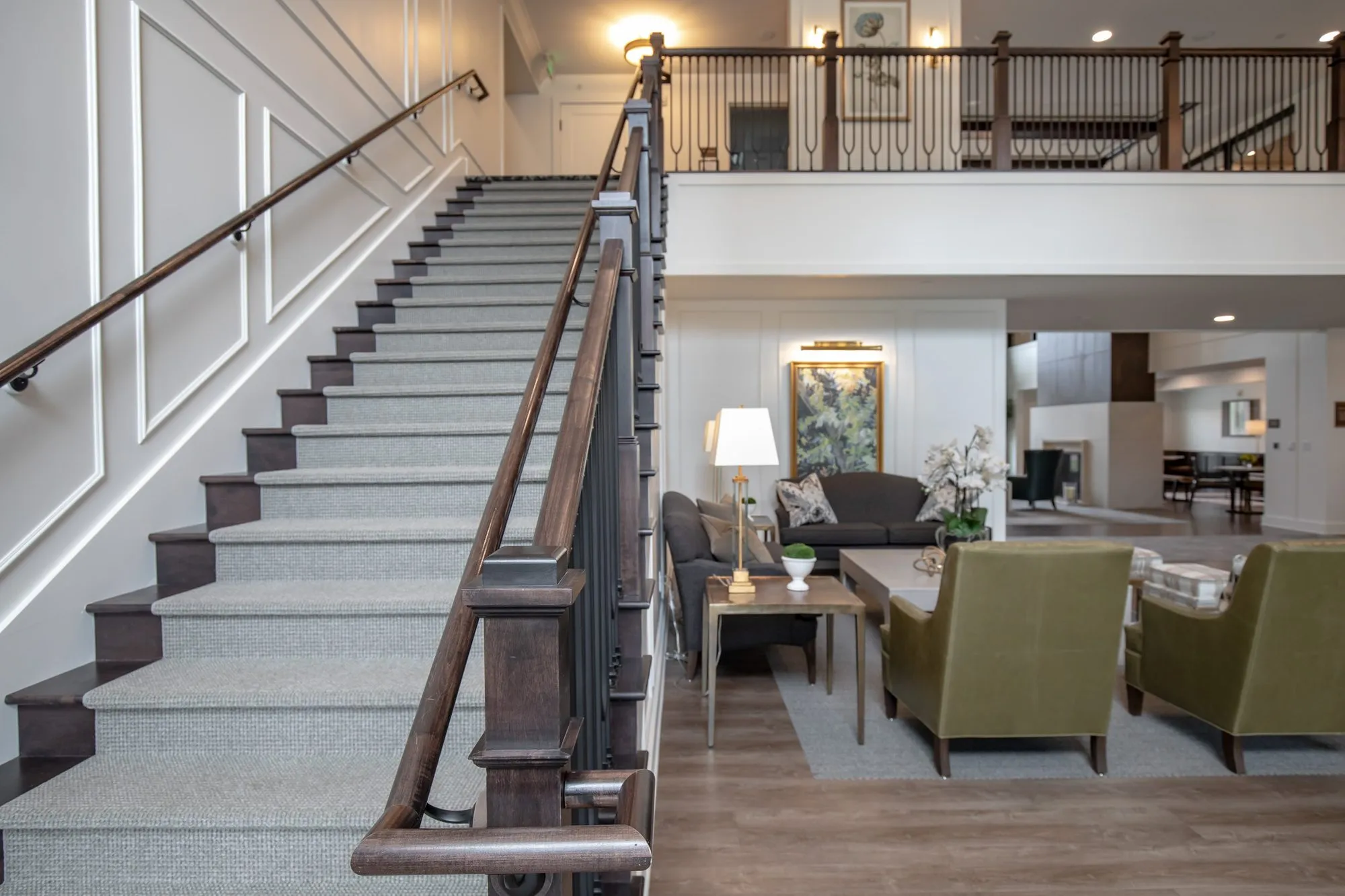 Active adult community stair with a dark wood handrail beside a living room of olive armchairs and a grey sofa