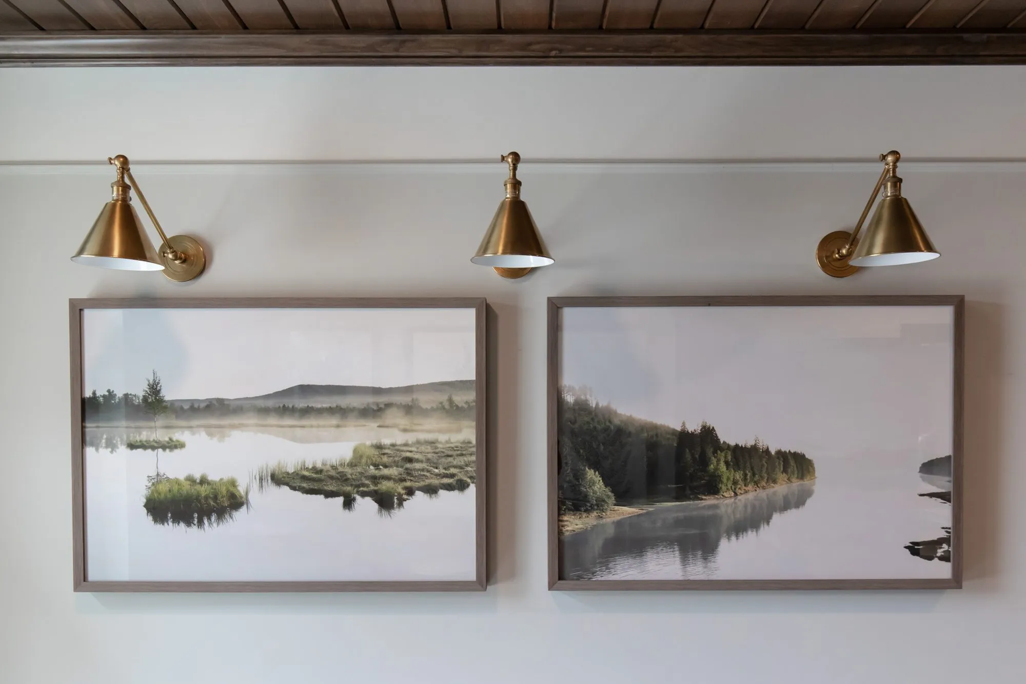 Active adult community interior detail: two framed misty lake photographs below three brass swing-arm sconces