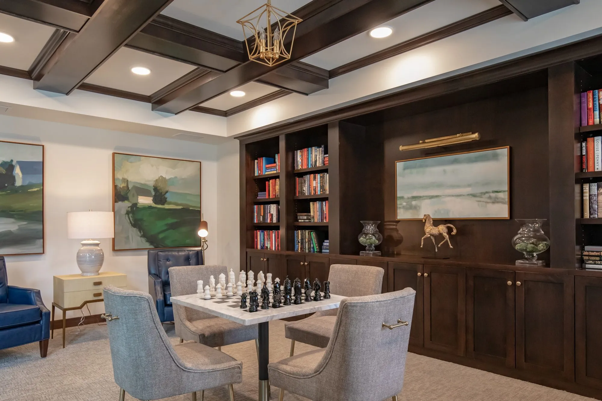 Active adult community library with a chess set on a game table, grey chairs and dark bookcases under a coffered ceiling