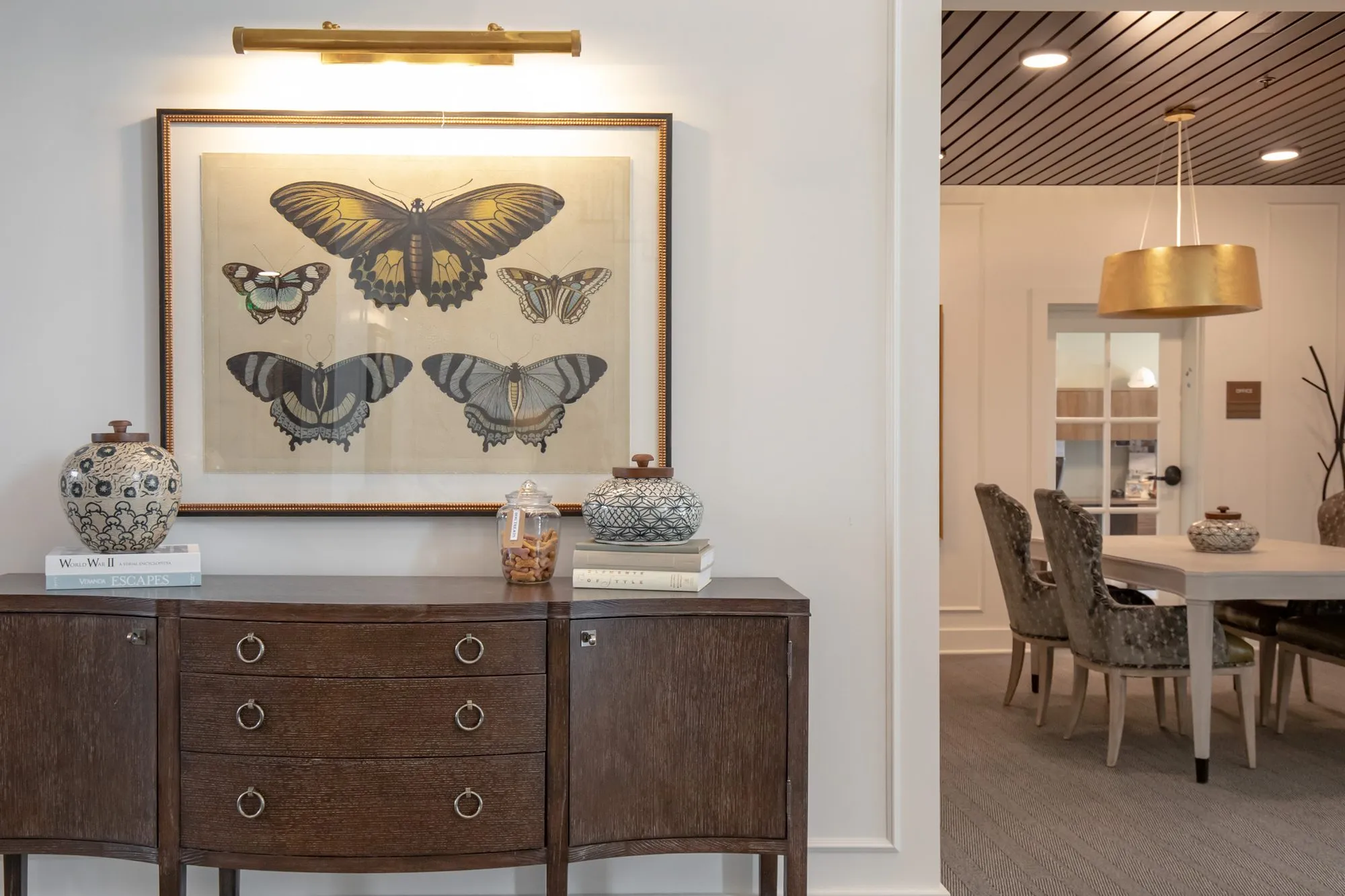 Active adult community interior detail: butterfly print over a dark sideboard, a small dining room with a gold pendant beyond