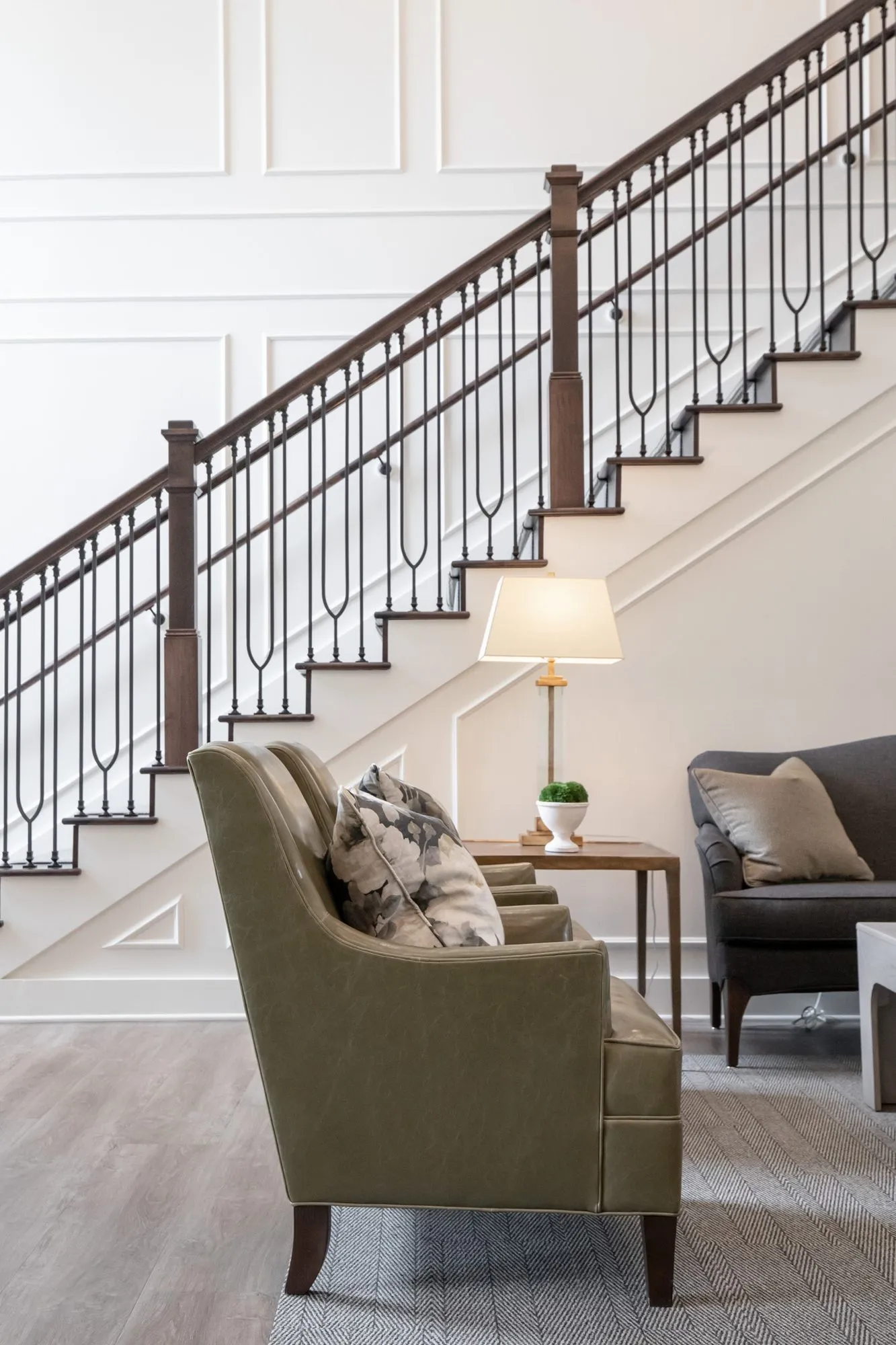 Active adult community interior detail: olive armchair and table lamp beneath a staircase with iron balusters