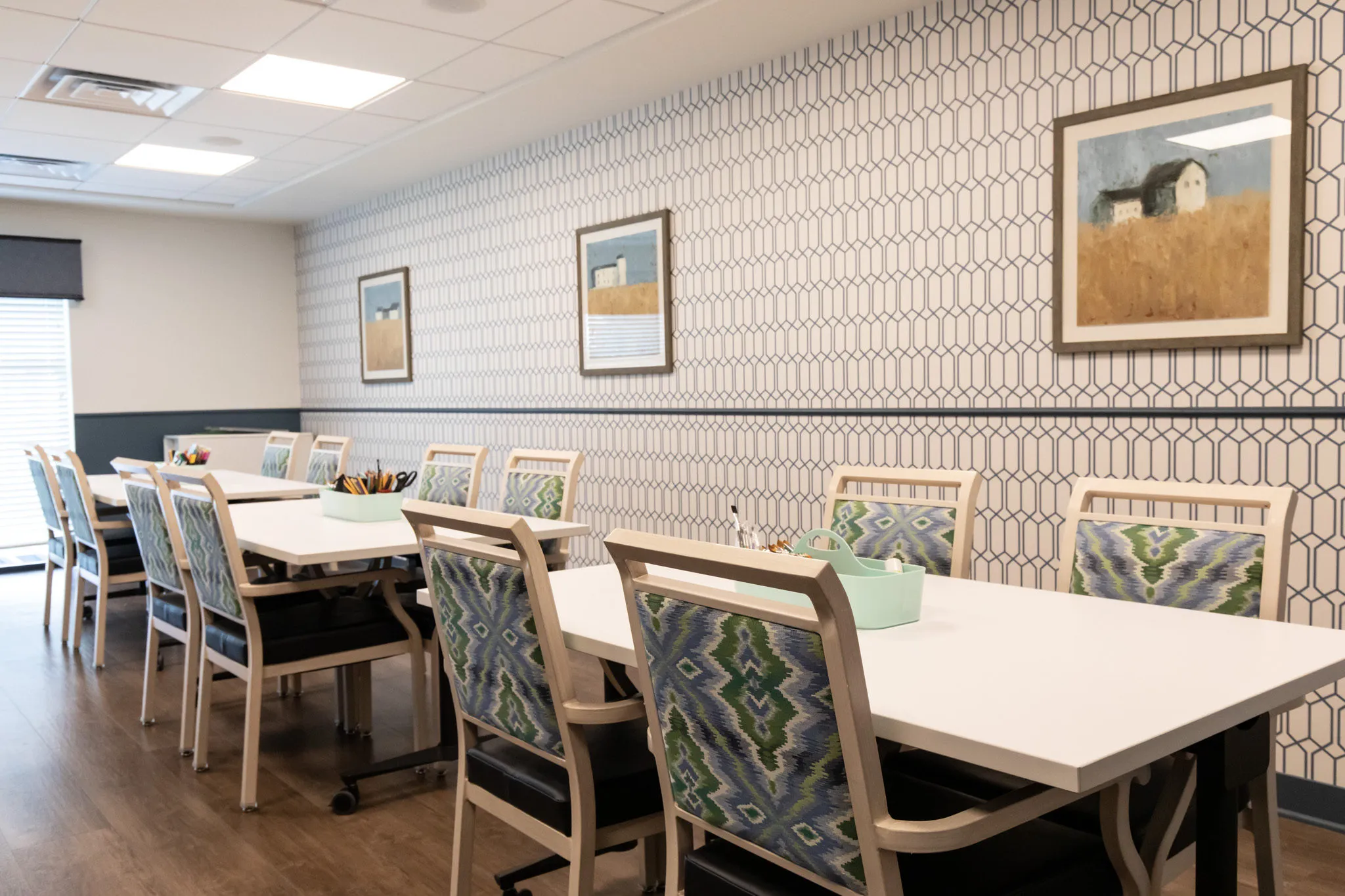 Active adult community activity room with long work tables and patterned chairs against geometric wallpaper and pastoral art