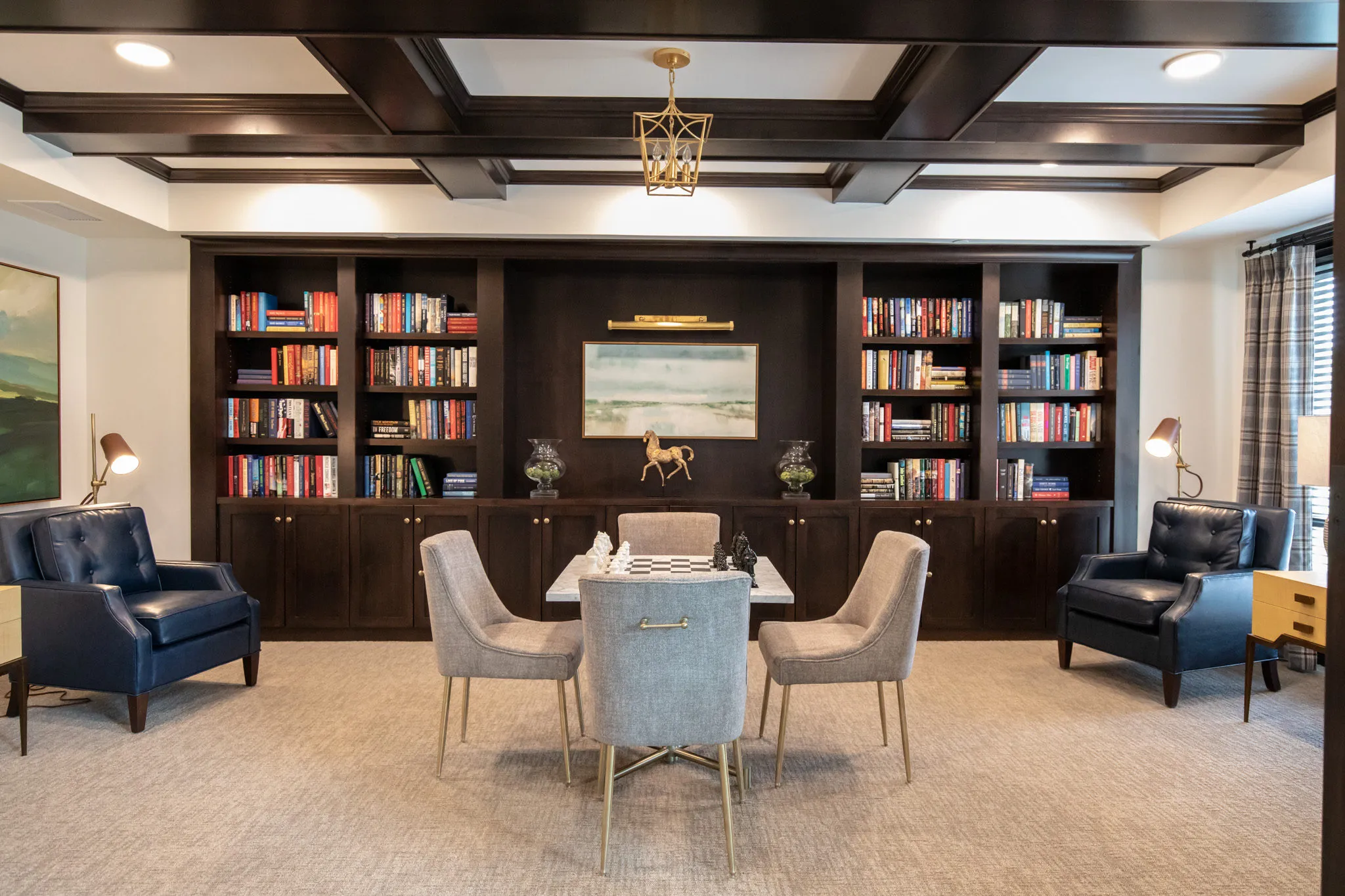 Active adult community library with dark built-in bookcases and a coffered ceiling around a card table set for four