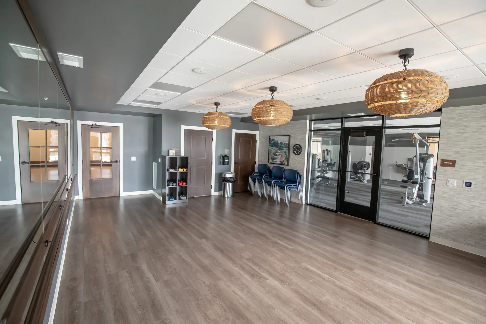 Active adult community yoga studio with a mirrored barre wall and woven pendants, the fitness room glazed beyond