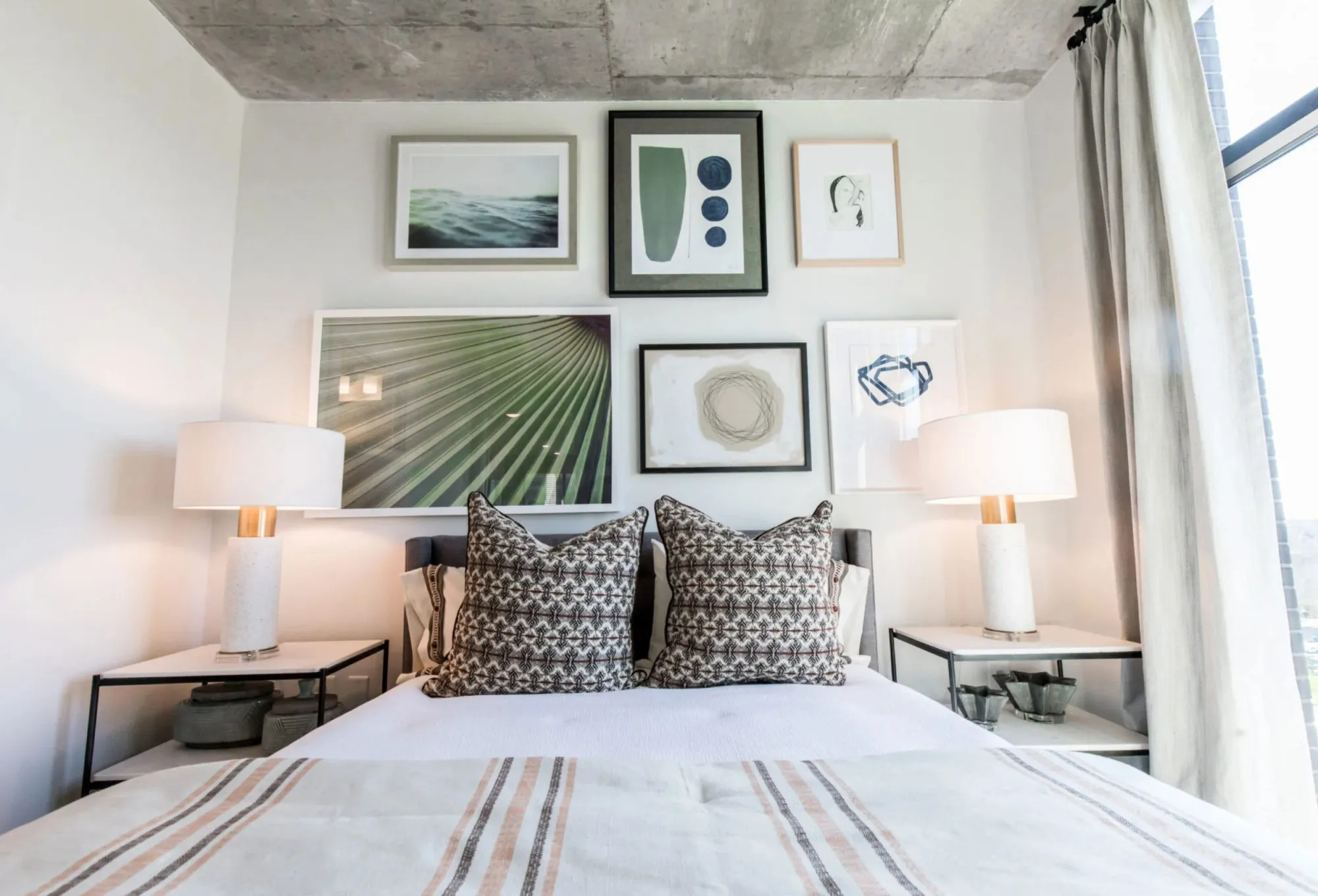 Multifamily apartment bedroom with an exposed concrete ceiling, a gallery wall of framed prints above the bed and white lamps on black nightstands