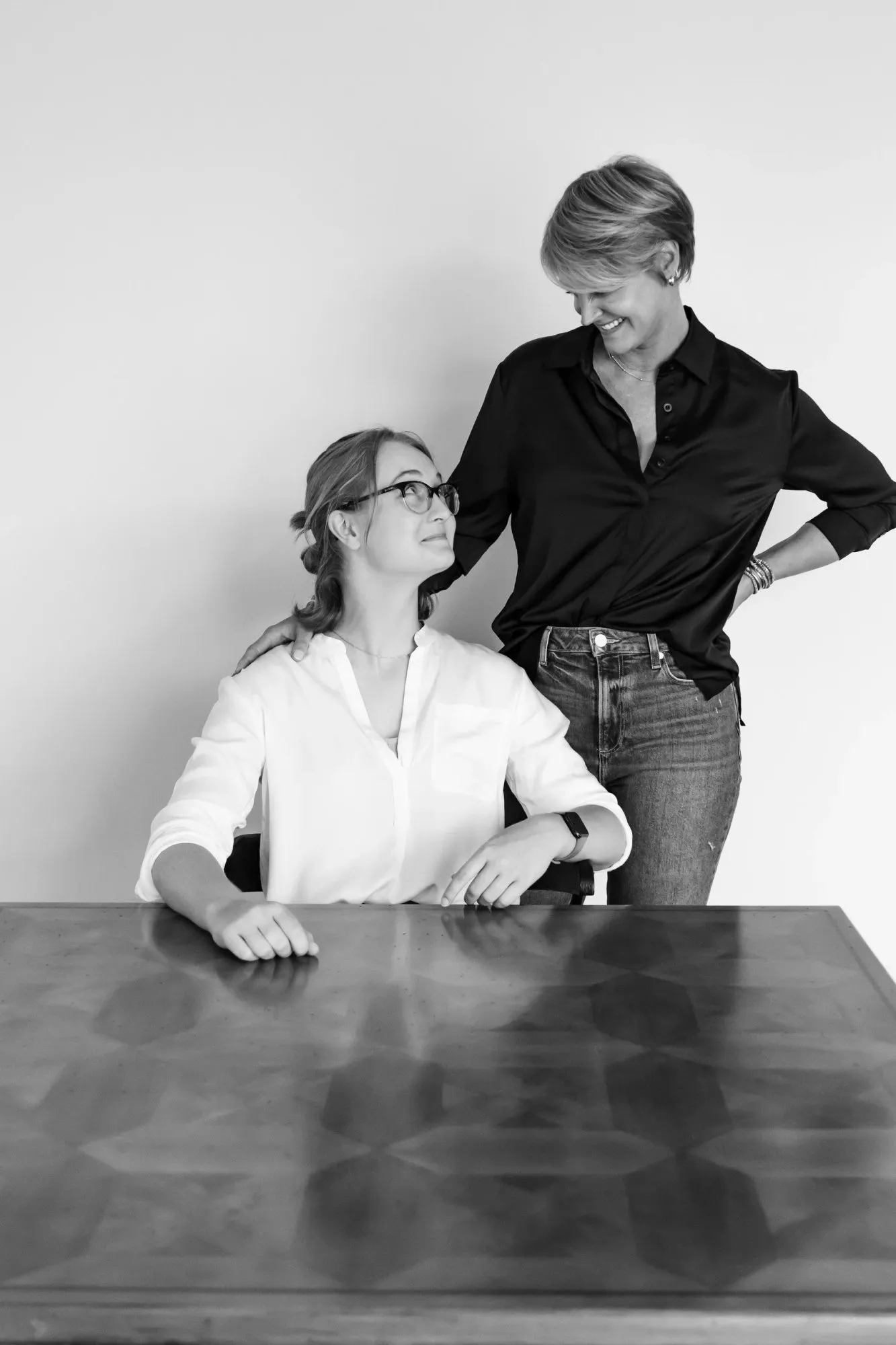 Two women in black and white: one seated at a wood table looks up at a colleague standing with a hand on her shoulder