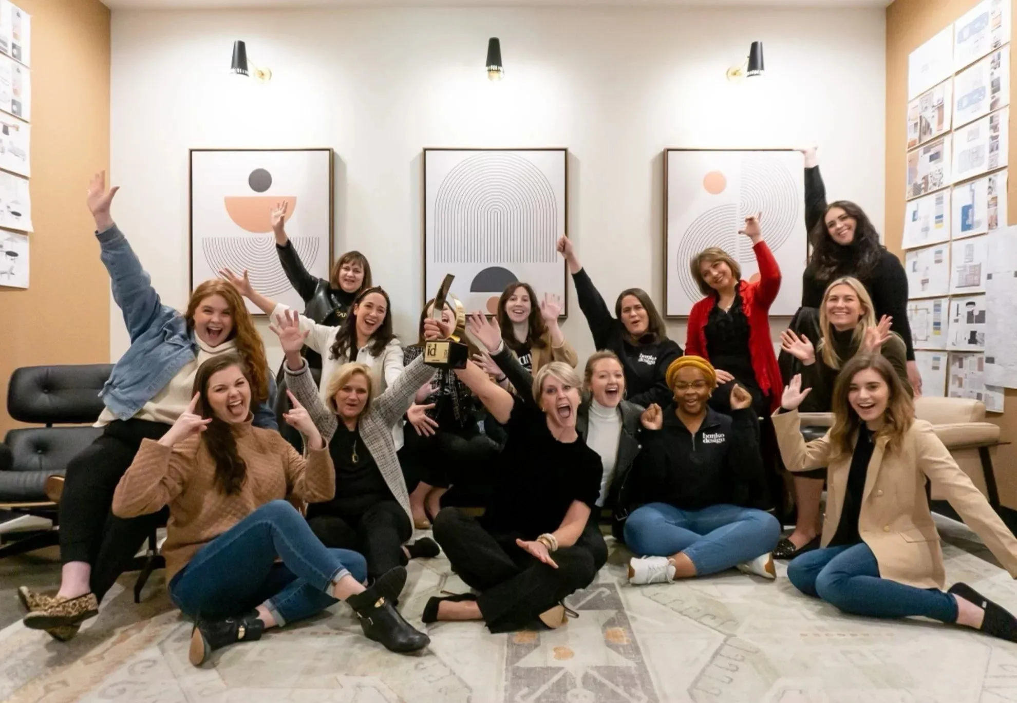 Banko Design staff seated on the studio floor with arms raised in celebration, one holding an award, framed prints on the wall behind
