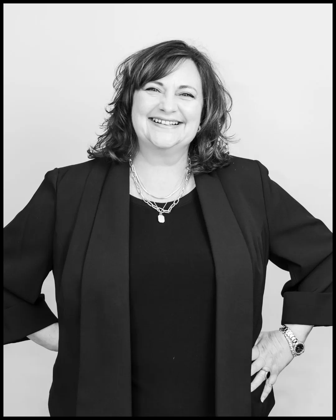 Betsy Distler in a black blazer with layered silver necklaces, hands on her hips, photographed in black and white