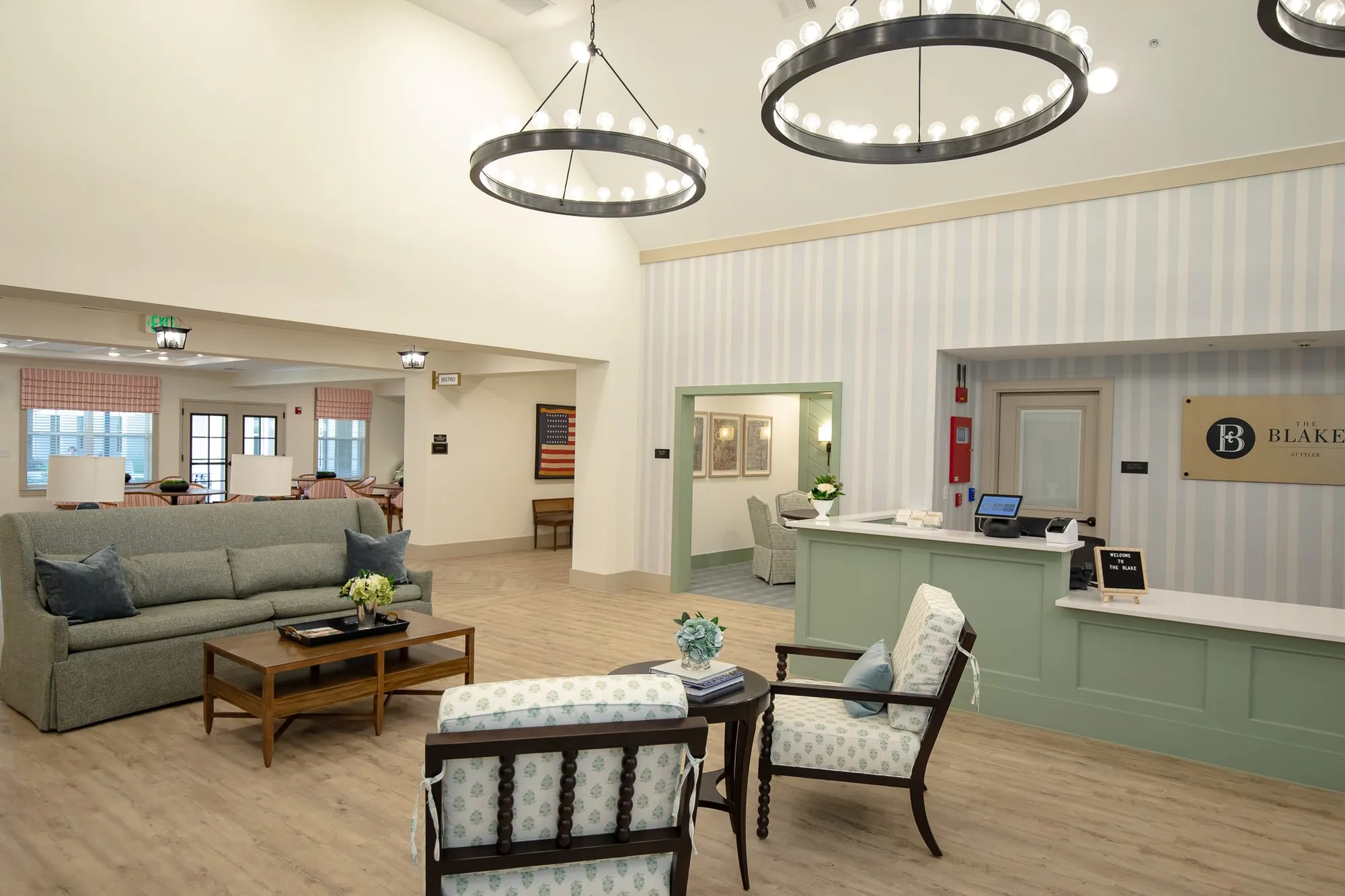 Senior living lobby under ring chandeliers, grey sofa and patterned armchairs, the sage reception desk at right