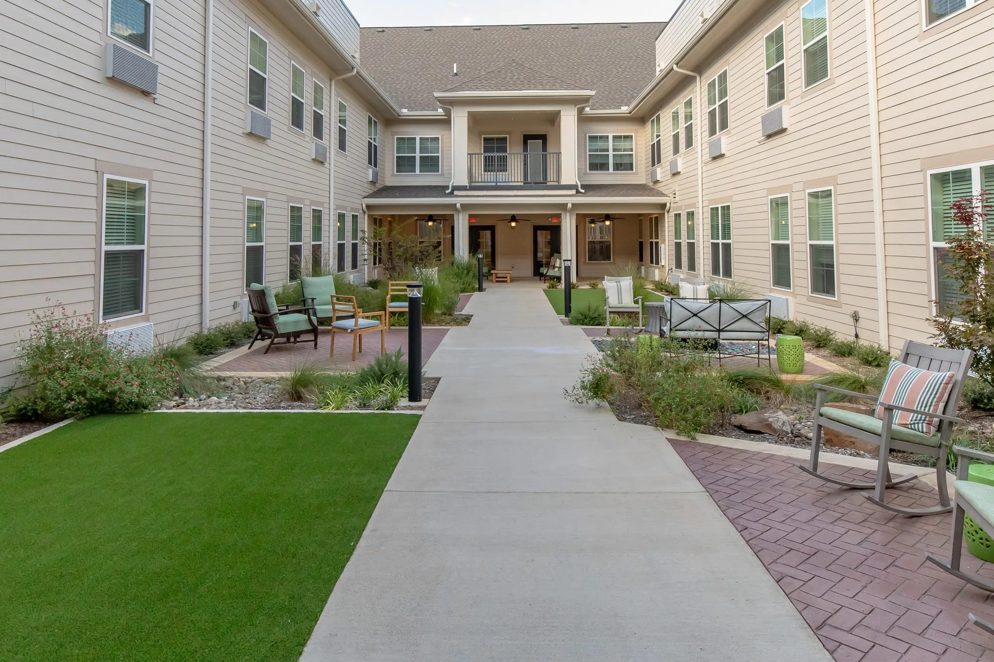 Senior living courtyard with a central walkway between turf and planting beds, rocking chairs and two storey wings