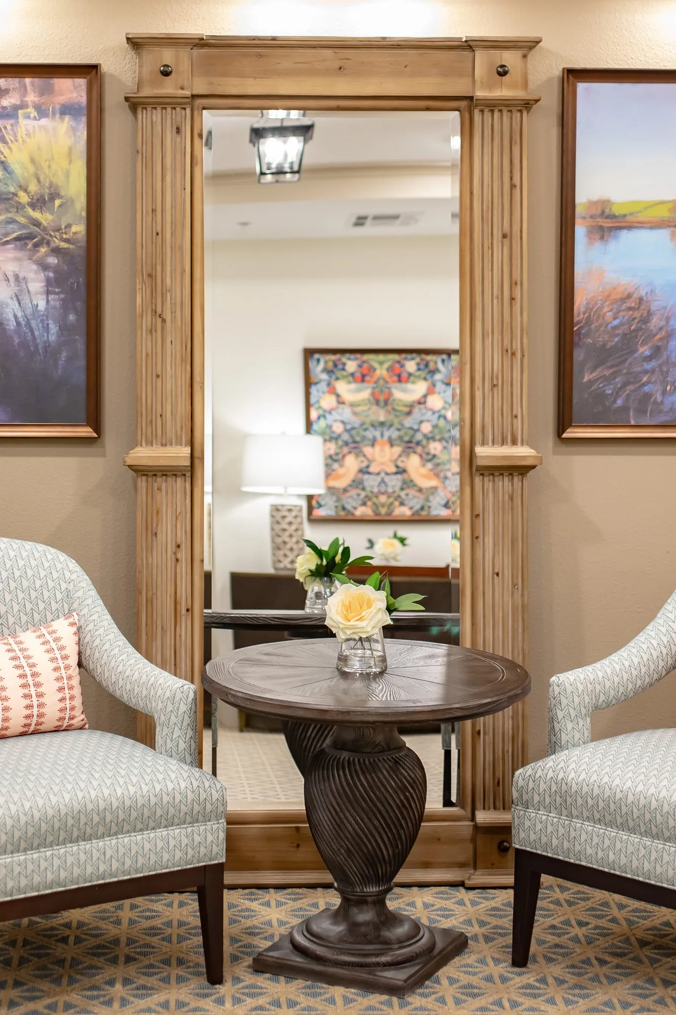 Senior living interior detail: patterned armchairs at a carved pedestal table before a wood framed mirror between landscapes