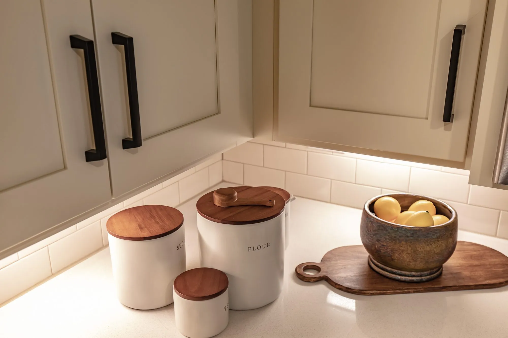 Senior living kitchen detail: white canisters and a wood bowl of lemons below taupe cabinets with black pulls
