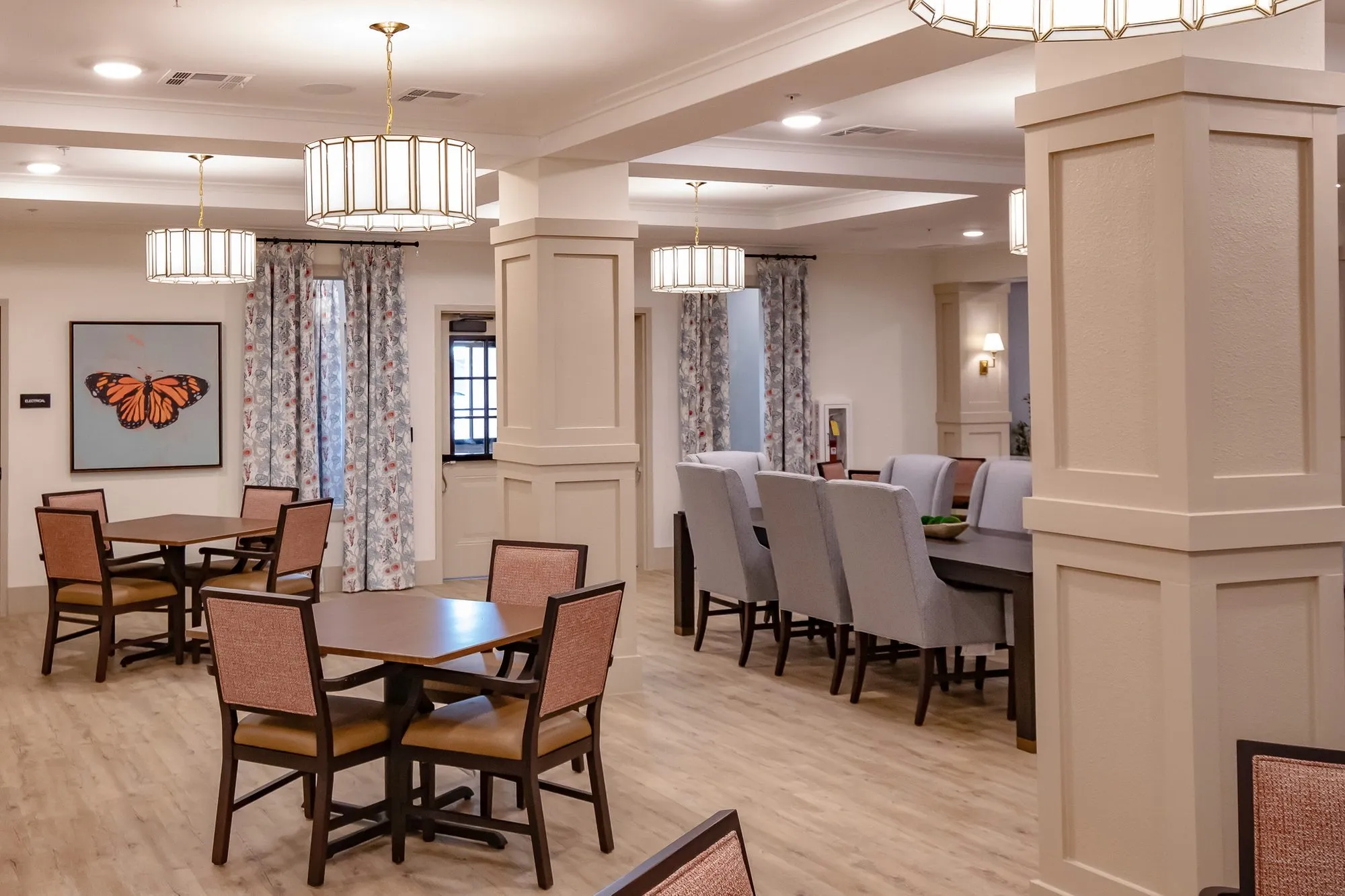 Senior living dining room with coral chairs at square tables, a butterfly painting and a long table of grey chairs beyond
