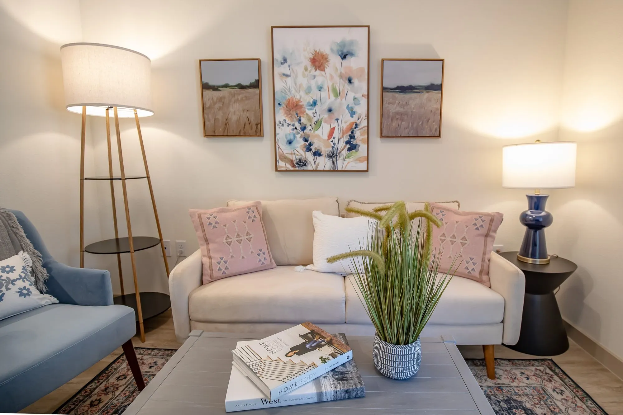Senior living model living room with a cream loveseat and blue armchair around a coffee table, a floral painting above