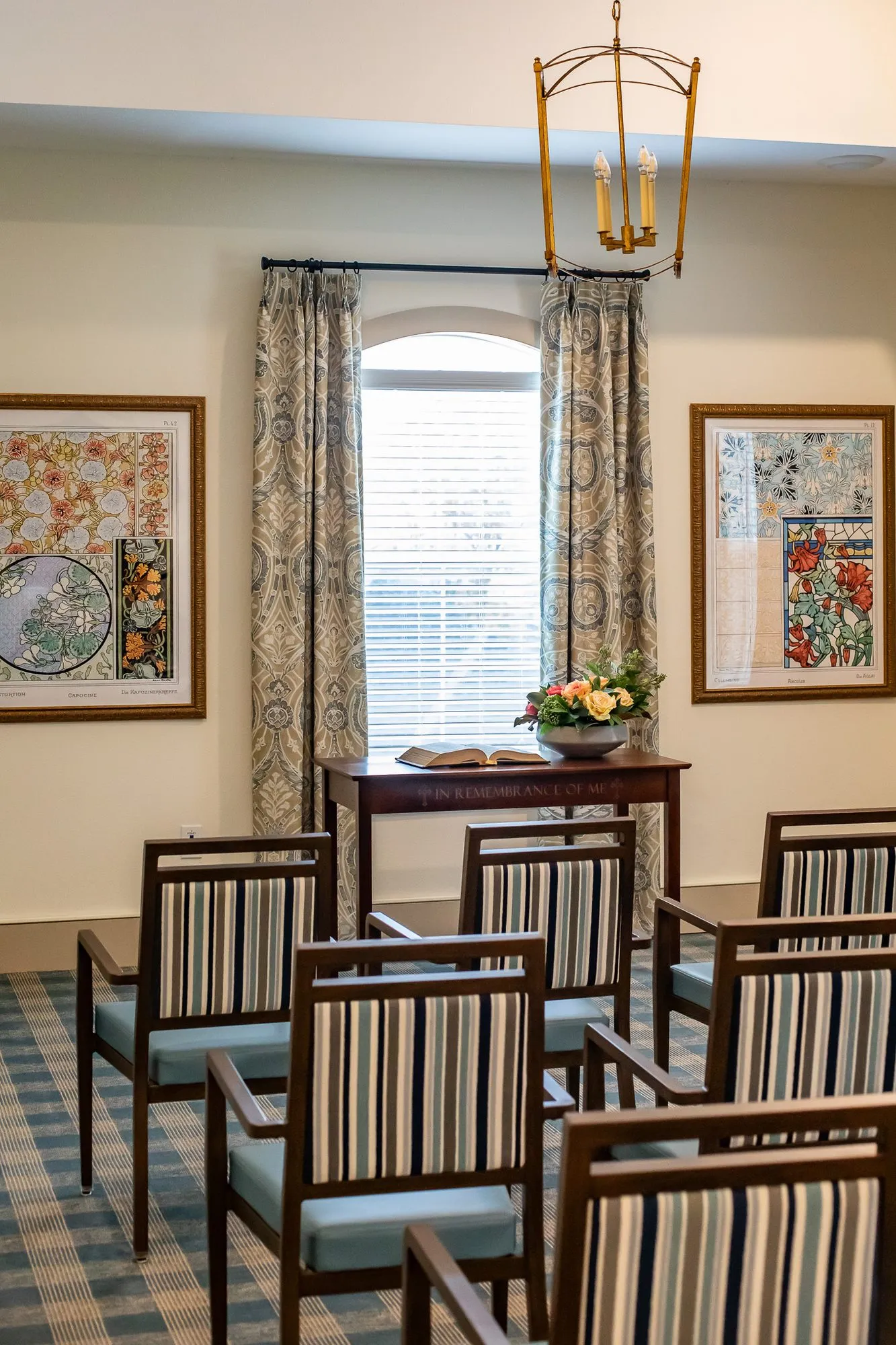 Senior living chapel with striped chairs facing a table with flowers under a brass lantern, framed floral panels