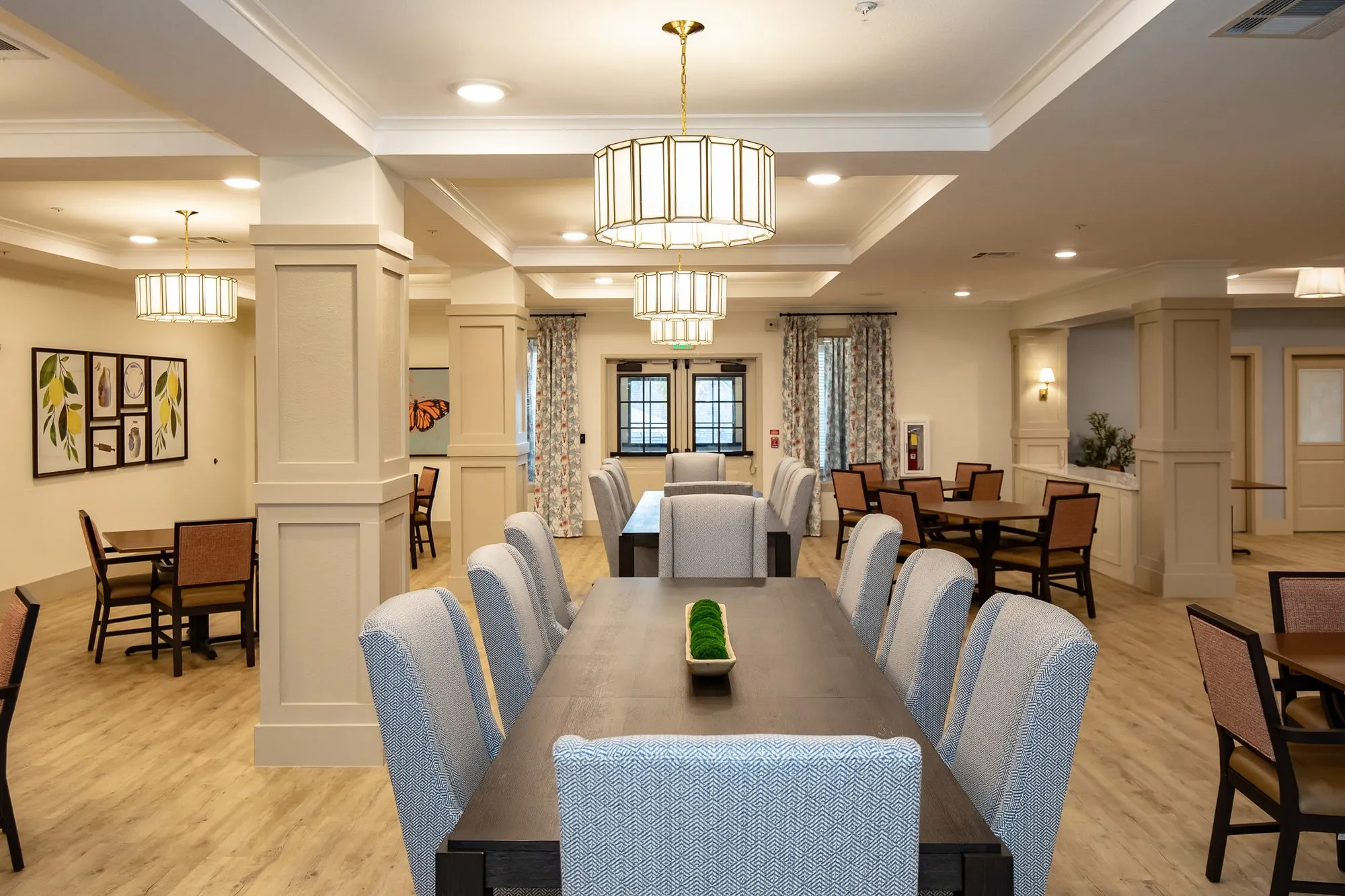 Senior living dining room looking down a long table of blue grey chairs under drum lantern pendants, columns either side