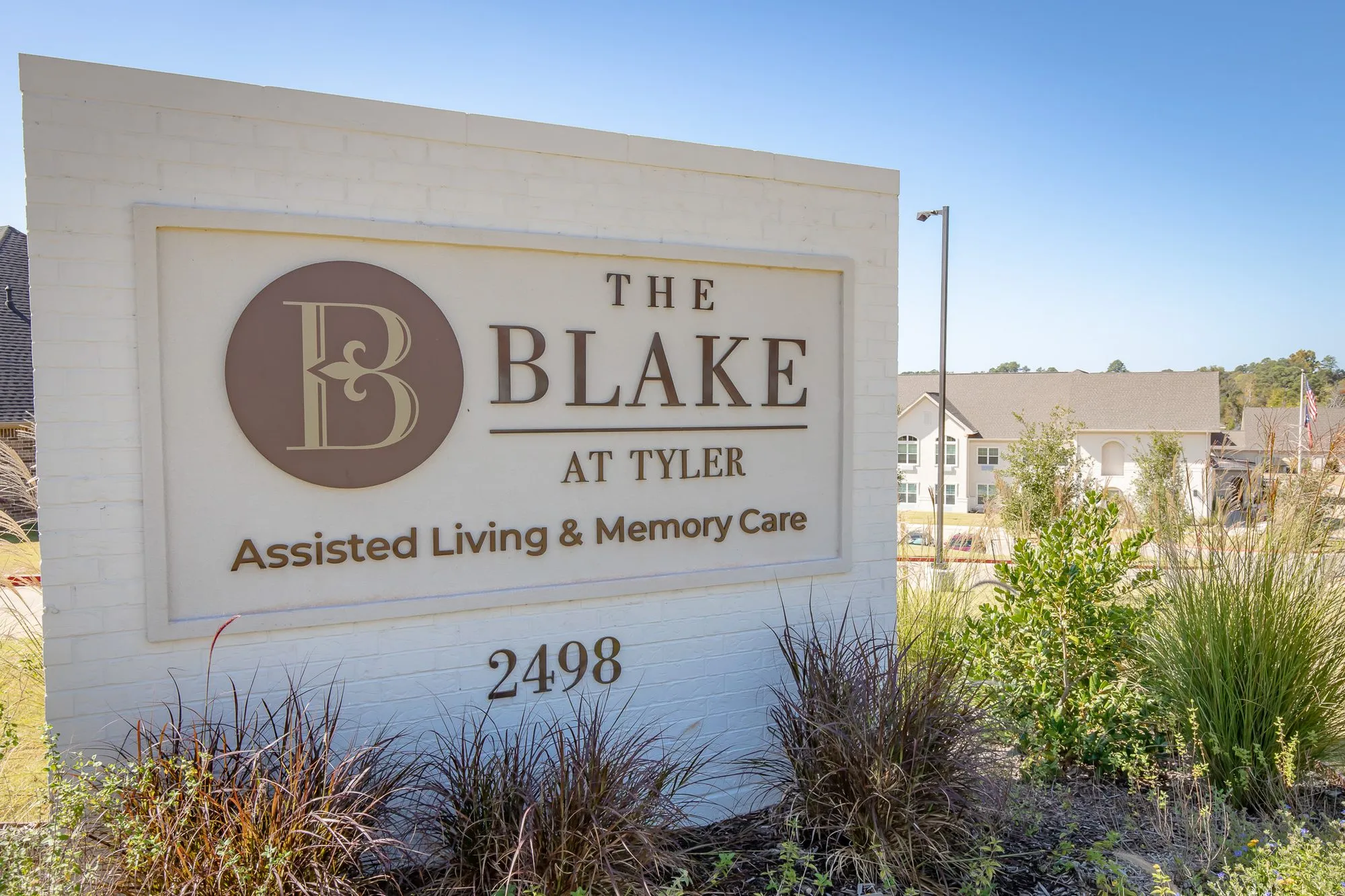 Senior living community exterior: monument sign on a white brick wall with a circular monogram and street number