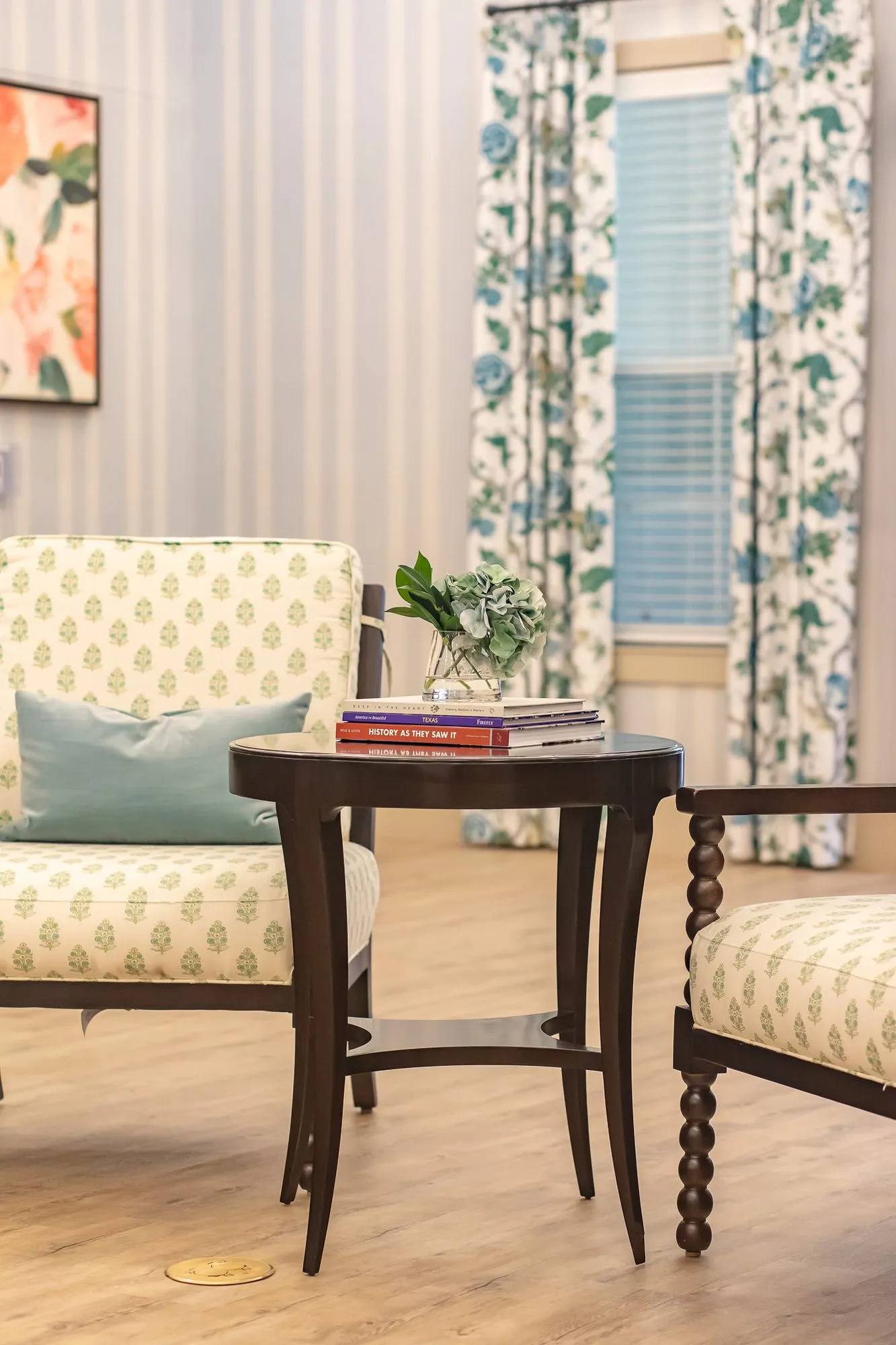 Senior living interior detail: block print armchairs beside a round side table with books and a plant, floral curtains