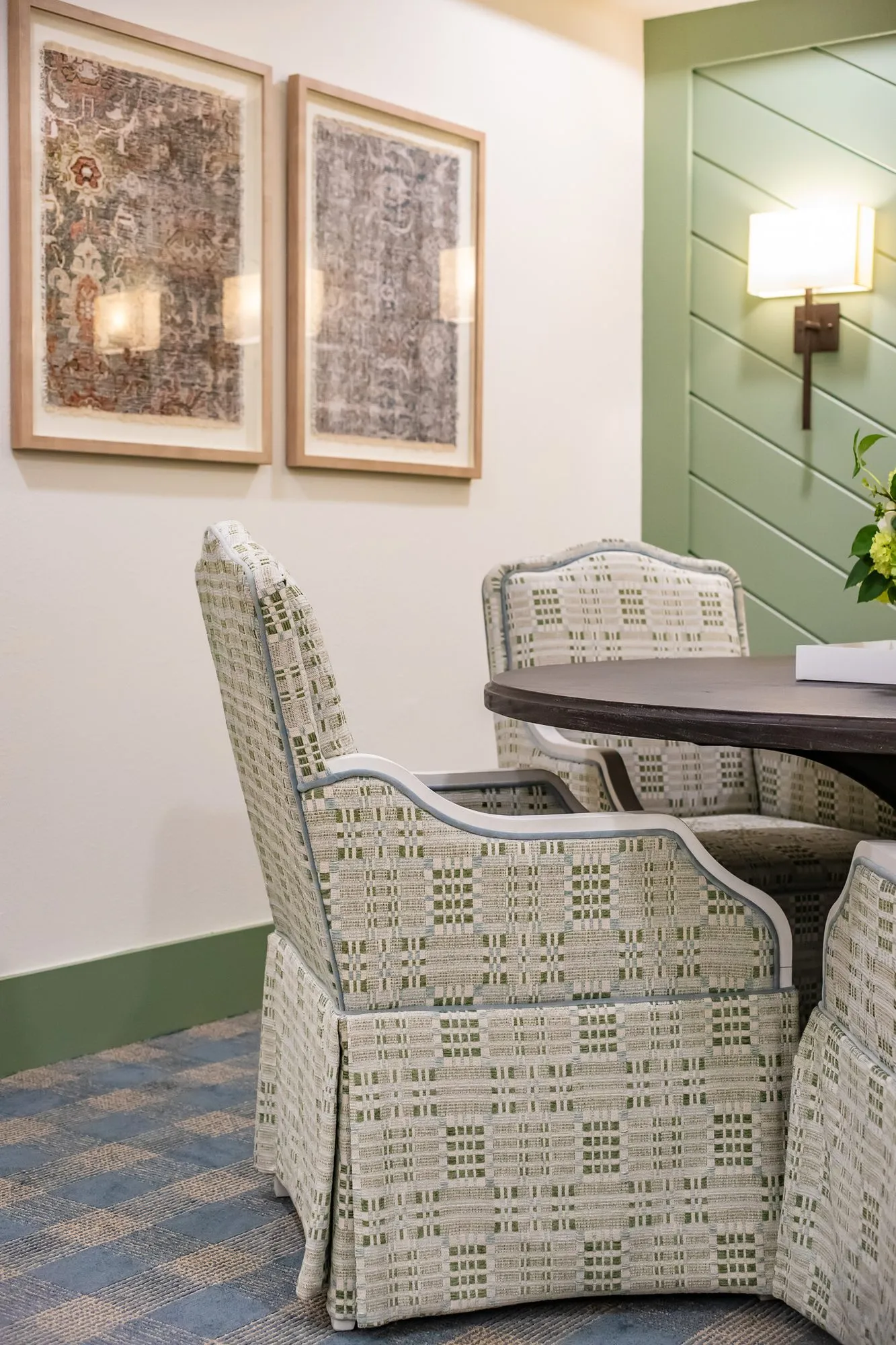 Senior living interior detail: patterned dining chairs at a round table below two textile prints, a sage shiplap wall beside