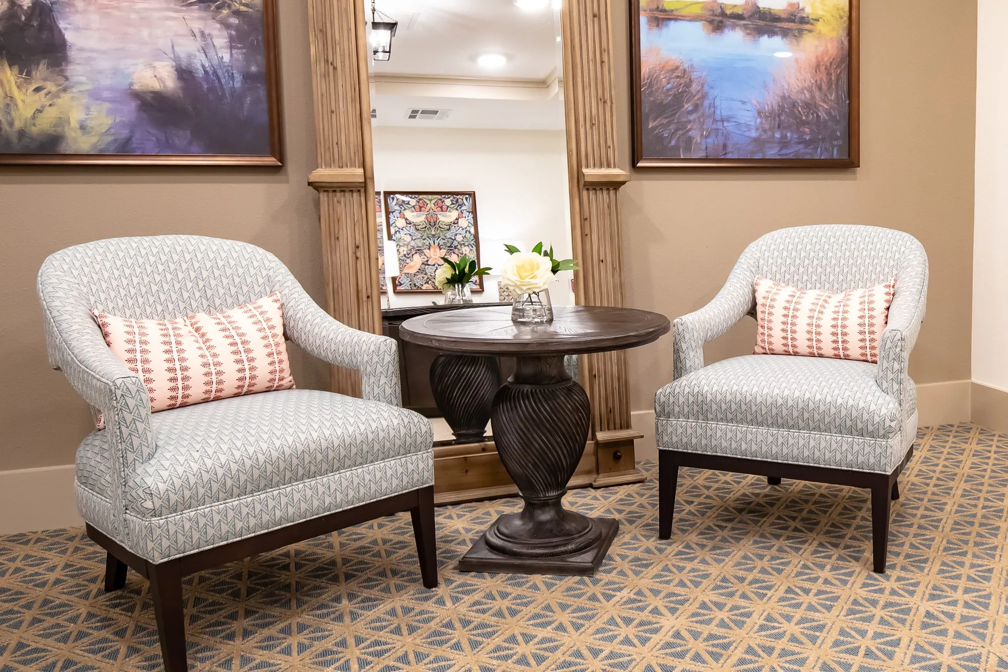 Senior living interior detail: two patterned armchairs with coral pillows at a carved pedestal table before a framed mirror