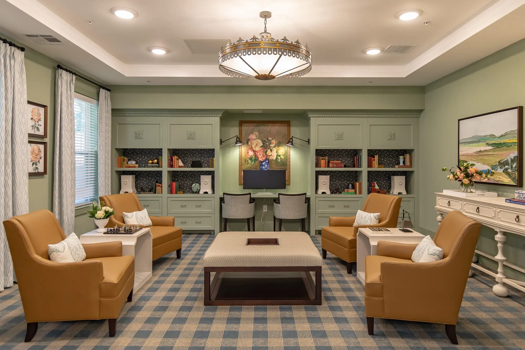 Senior living club room with camel leather chairs around a linen ottoman, sage built-ins framing two desk chairs beyond