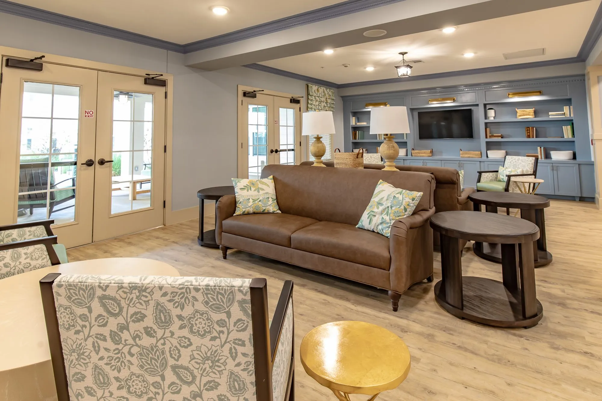 Senior living lounge with a brown leather sofa and round wood tables before blue grey built-ins, French doors at left