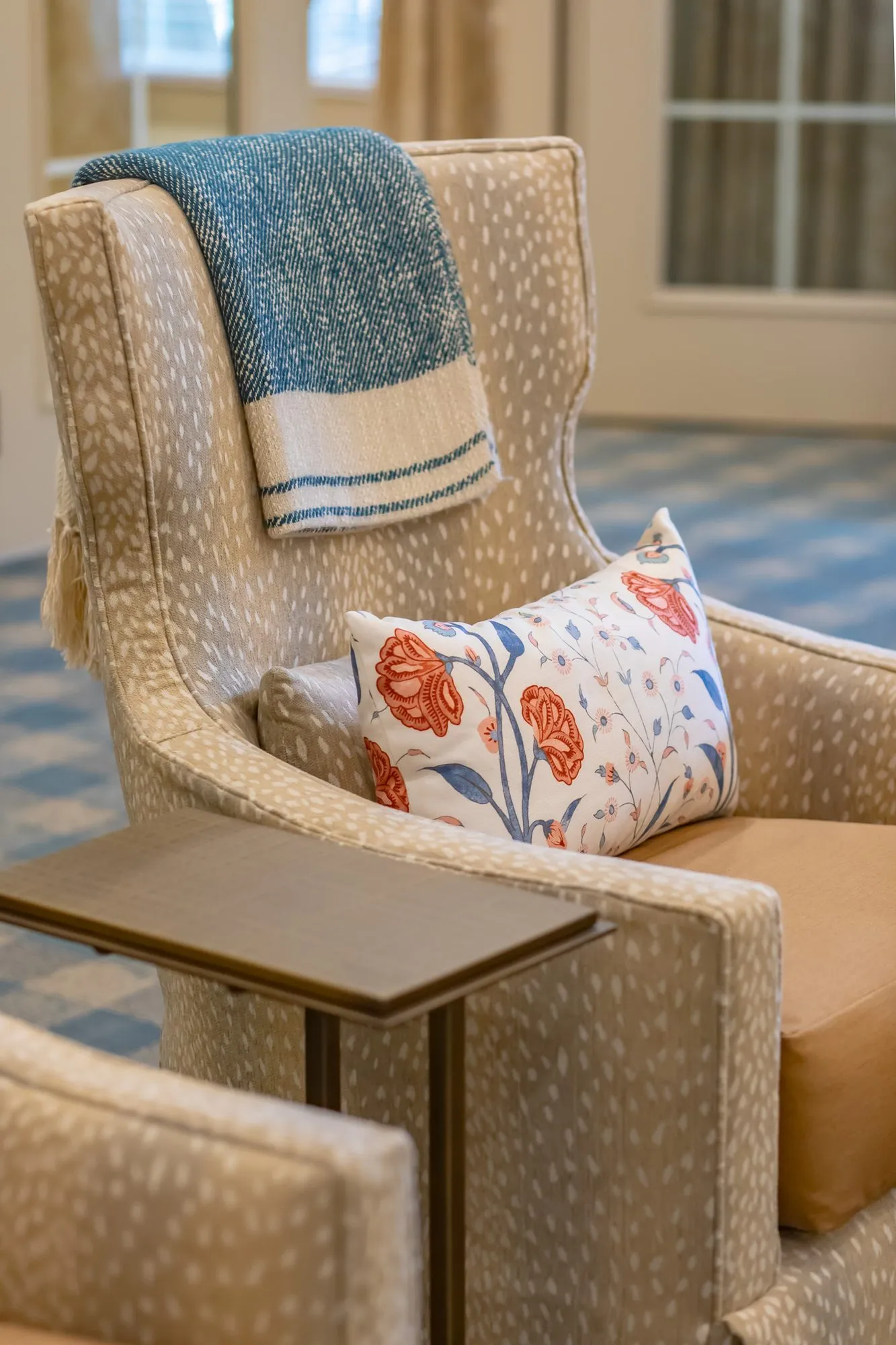 Senior living interior detail: fawn print wingback recliner with a blue throw and floral pillow beside a wood tray table