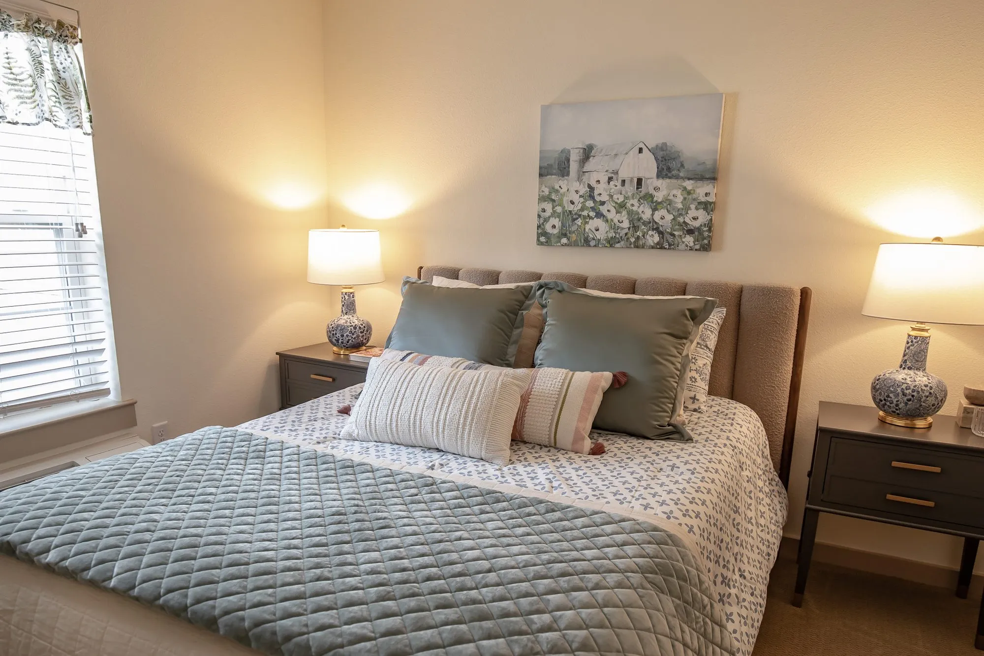 Senior living bedroom with sage velvet shams and a quilted blue throw on a tufted bed between two ginger jar lamps