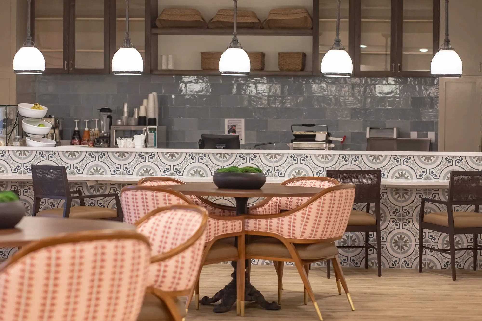 Senior living interior detail: coral striped bistro chairs at a pedestal table, glass pendants over the tiled counter