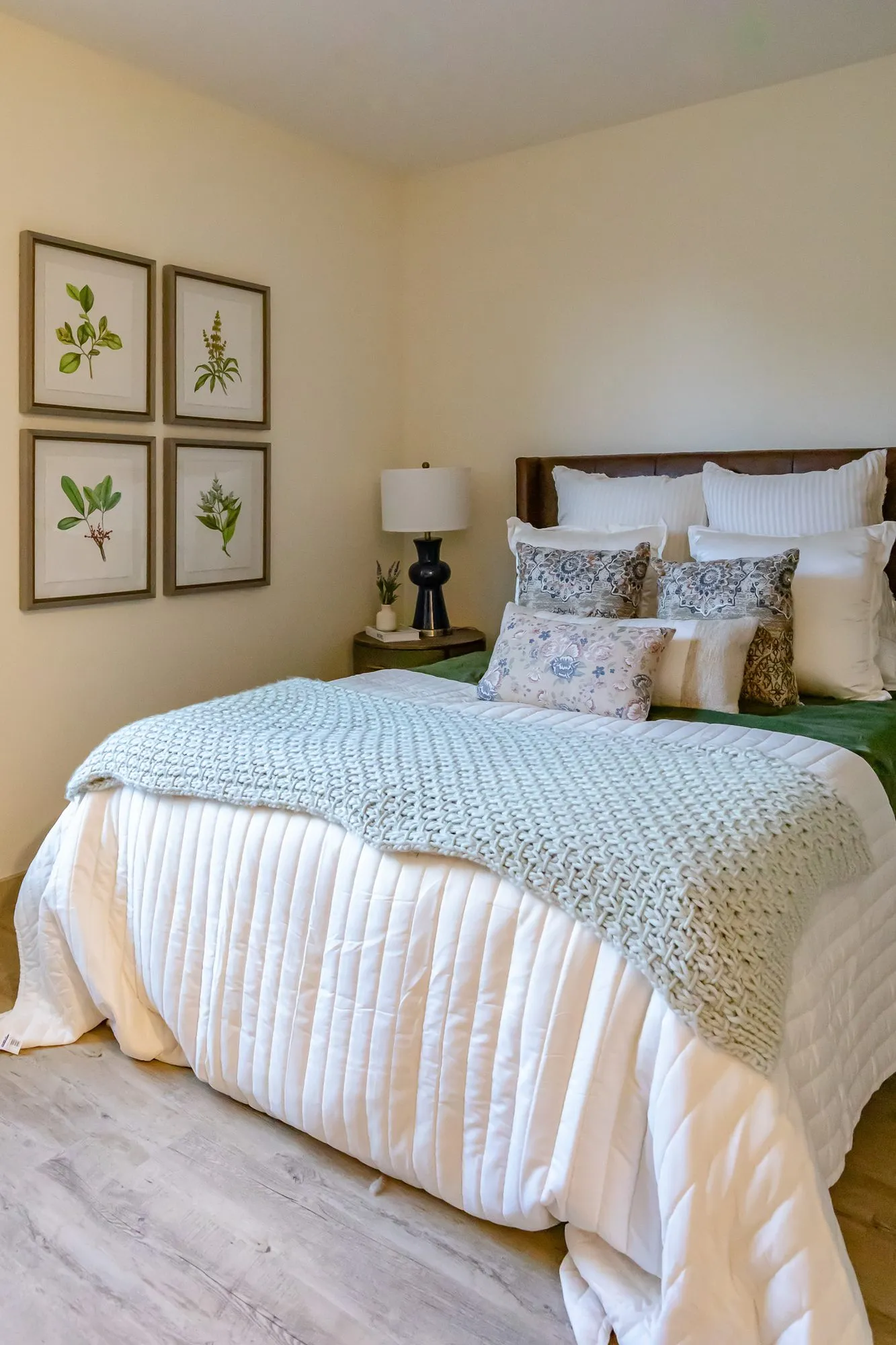 Senior living model bedroom with a leather headboard, white coverlet and blue knit throw below four botanical prints