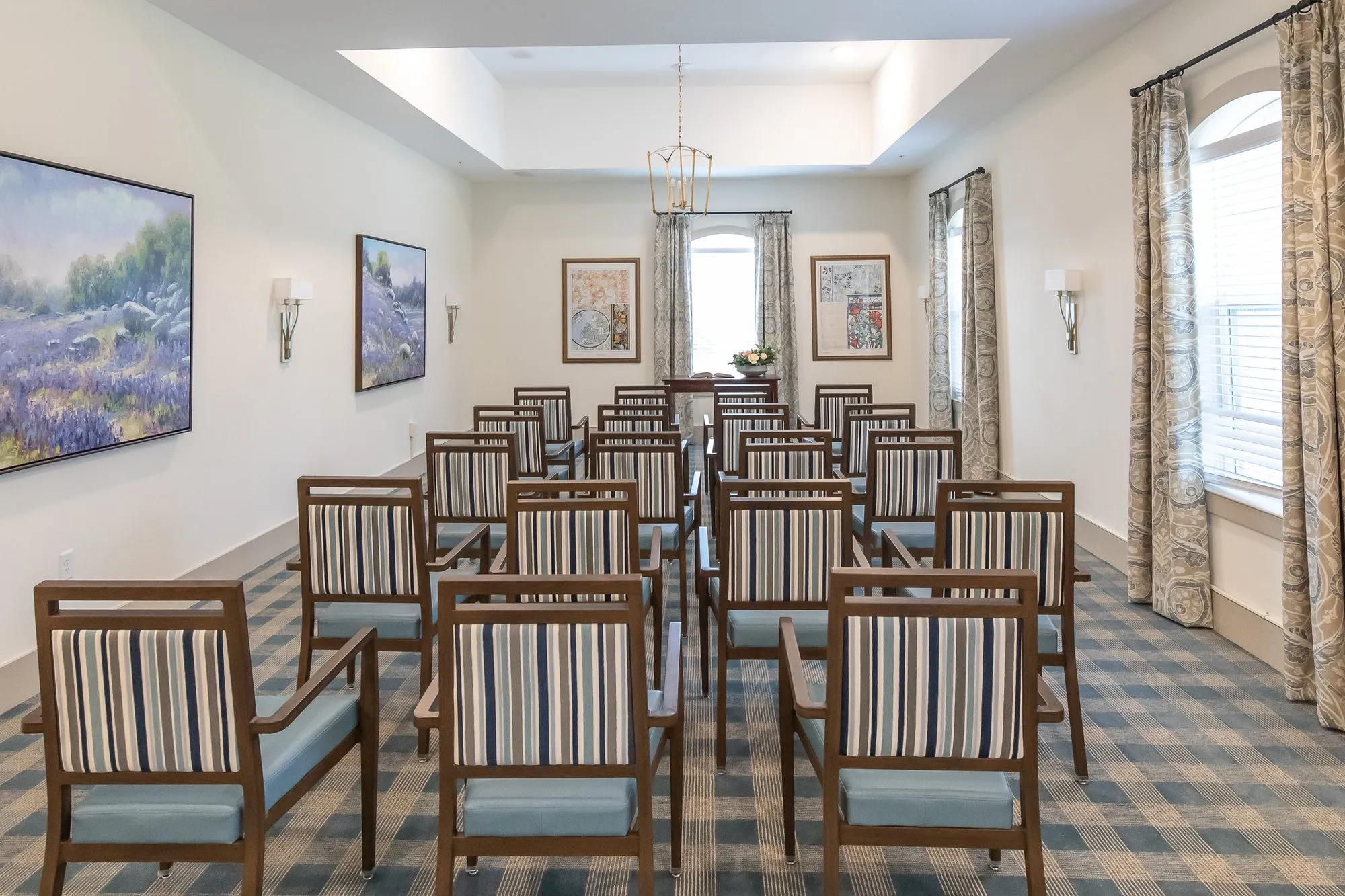 Senior living chapel from the back corner, rows of striped chairs on a plaid carpet, landscape paintings and arched windows