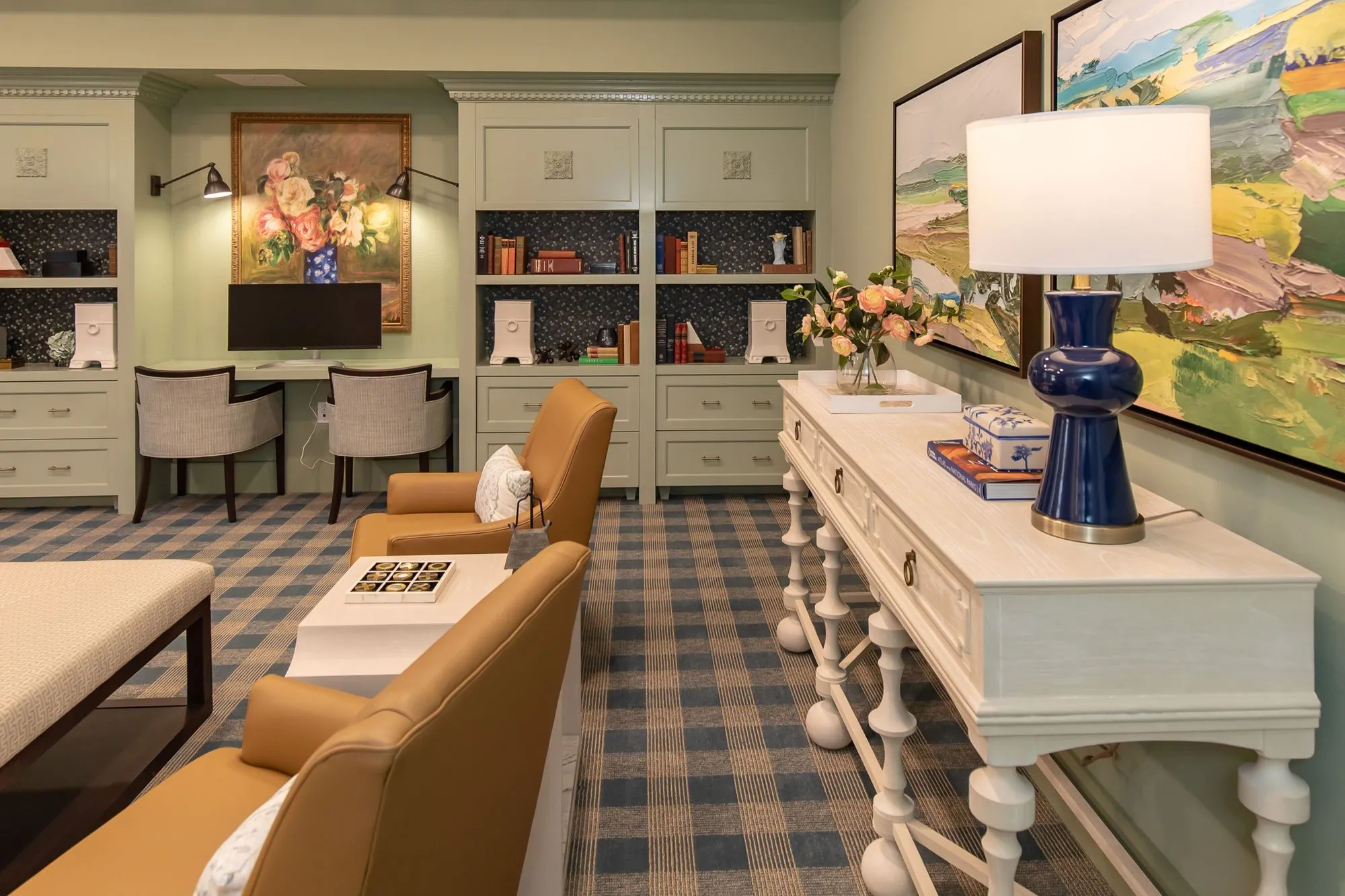 Senior living club room from the console with a navy lamp and landscape painting, camel chairs and sage built-ins beyond