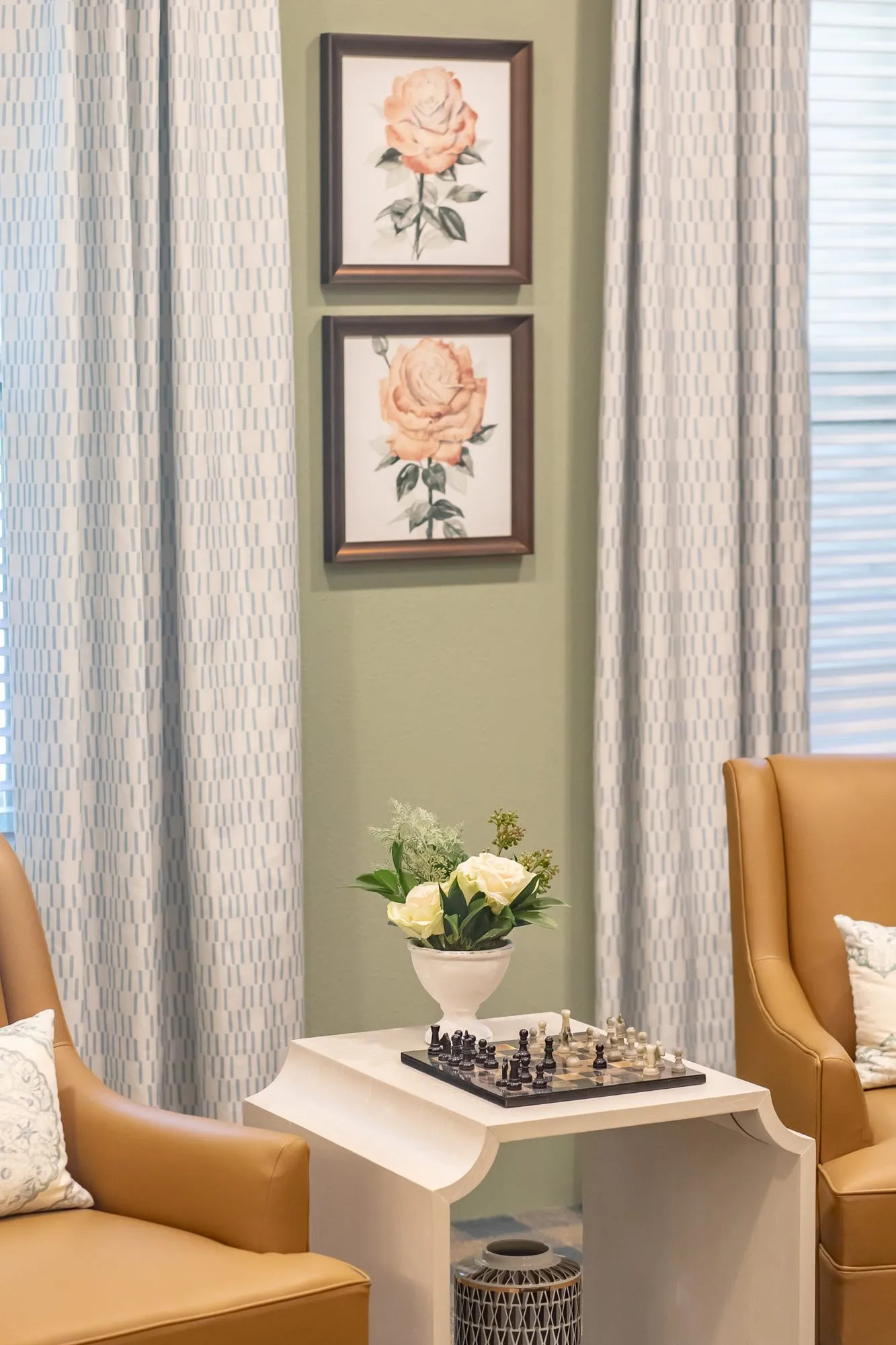 Senior living interior detail: chess set on a white side table between camel leather chairs below two rose prints