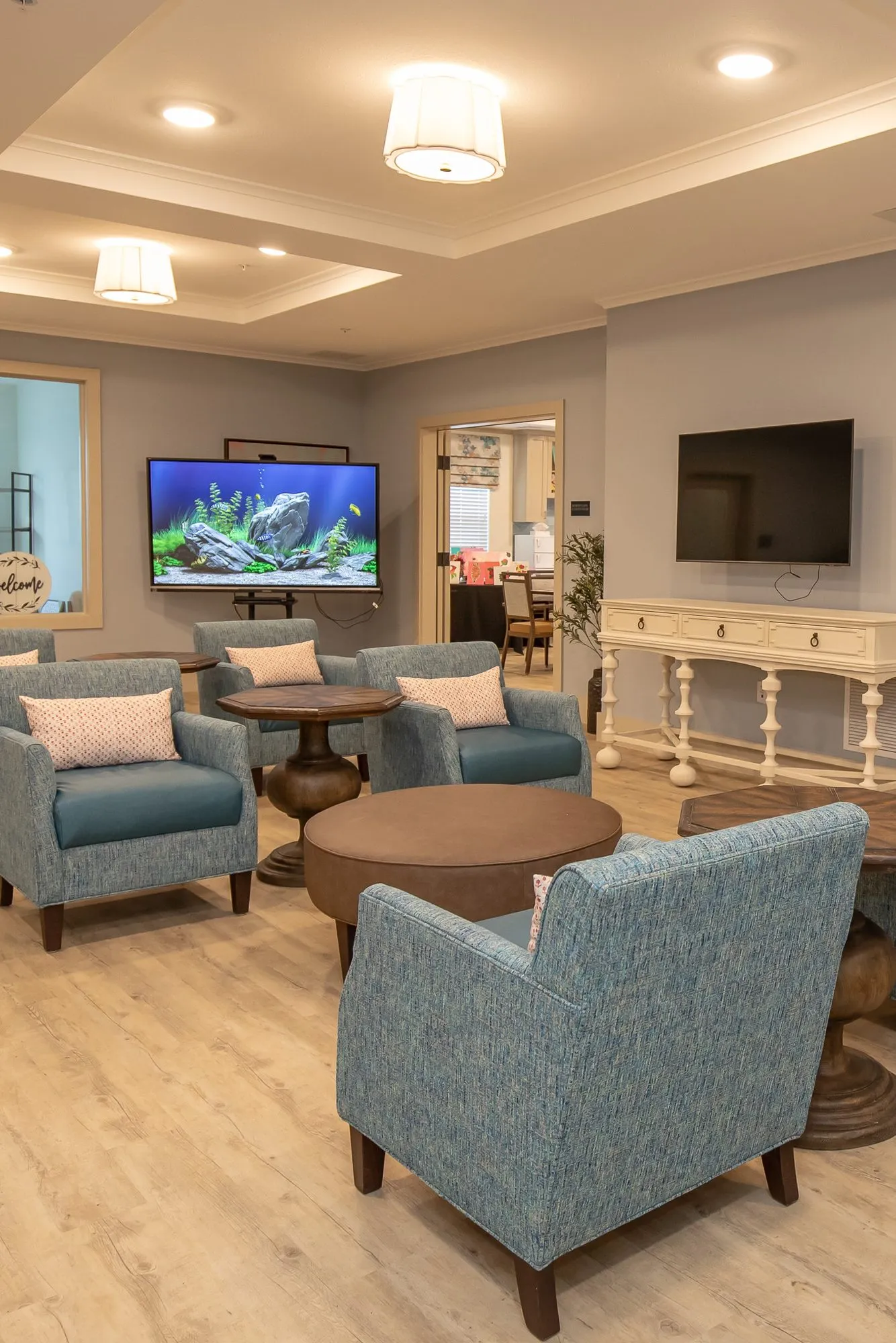 Senior living lounge with teal armchairs around round wood tables, a TV showing an aquarium scene and a white console