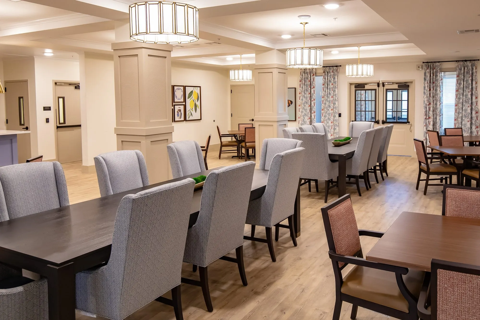 Senior living dining room across a long table with grey chairs toward coral chairs at square tables and curtained windows