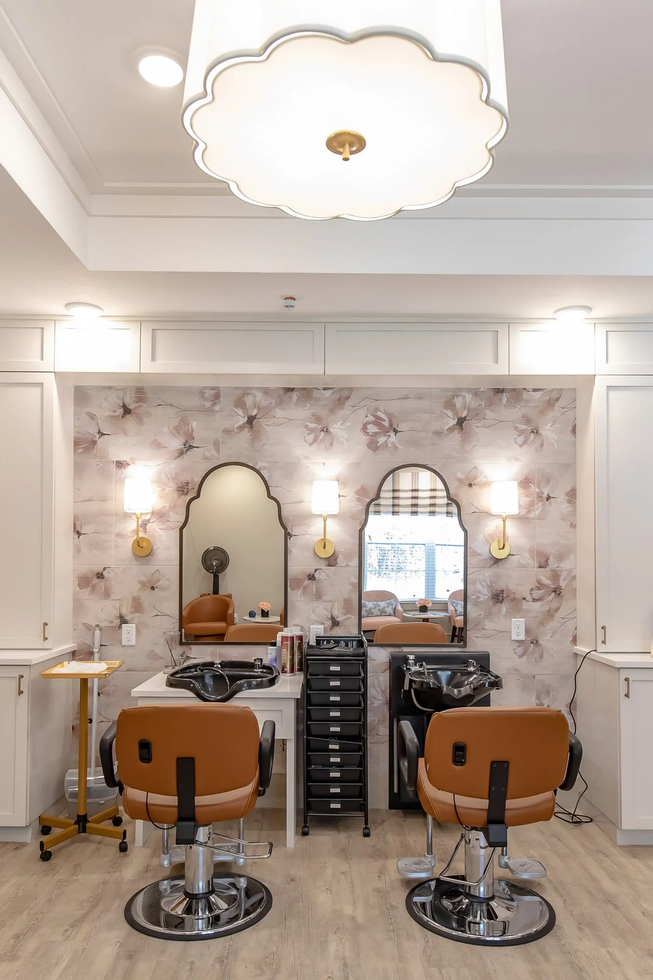 Senior living salon with two cognac styling chairs before arched mirrors on blush wallcovering under a scalloped light