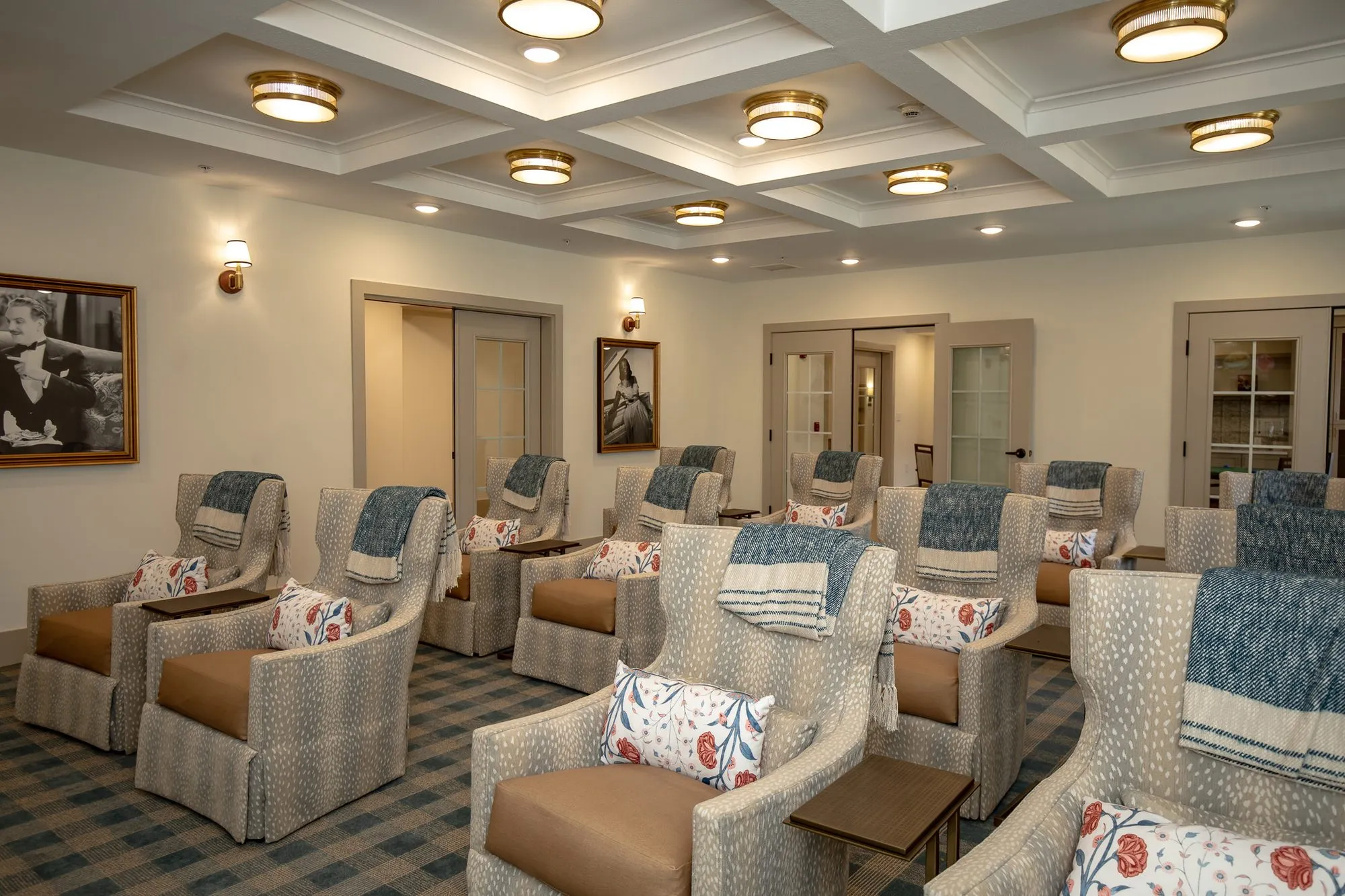 Senior living theater with rows of fawn print recliners and blue throws below black and white portraits, a coffered ceiling