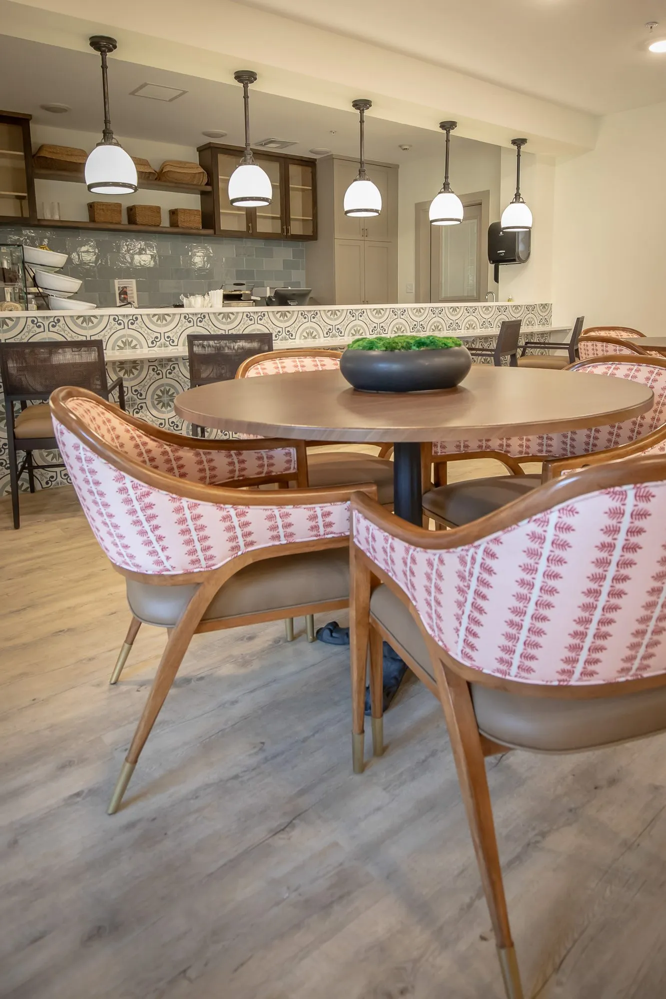 Senior living bistro with coral striped chairs at a round table before the tiled servery and five pendants