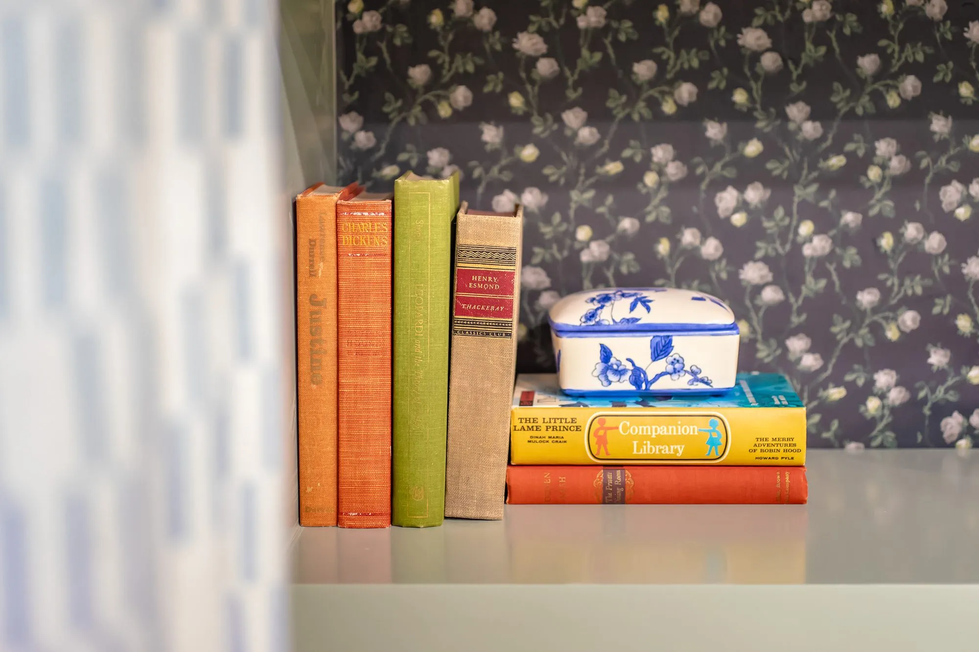 Senior living interior detail: orange, green and red vintage books and a blue and white lidded box on dark rose wallpaper