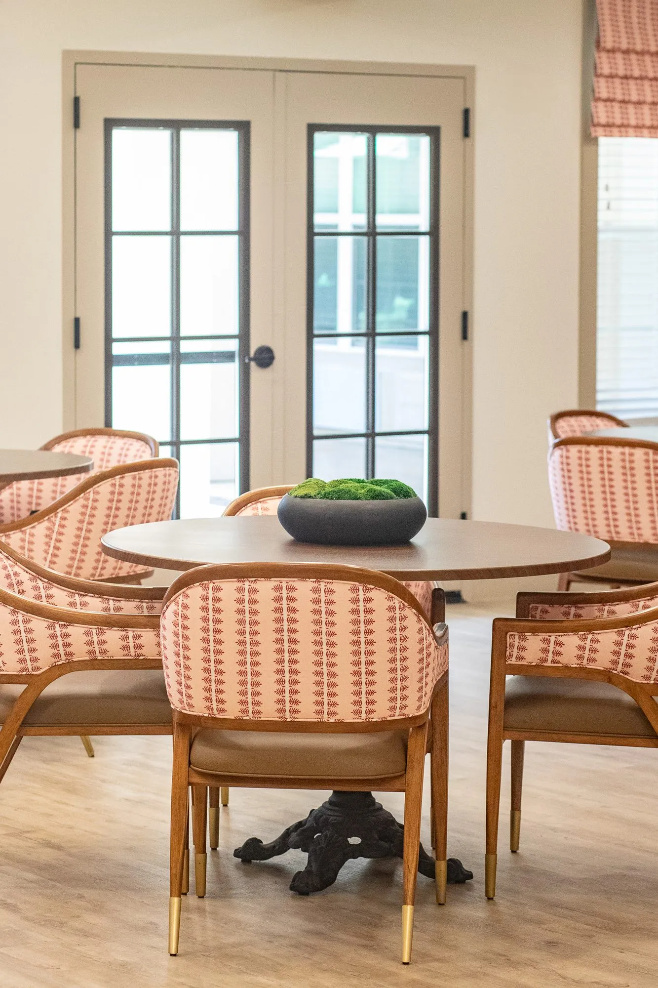 Senior living interior detail: coral striped chairs at a round pedestal table with a moss bowl, French doors behind