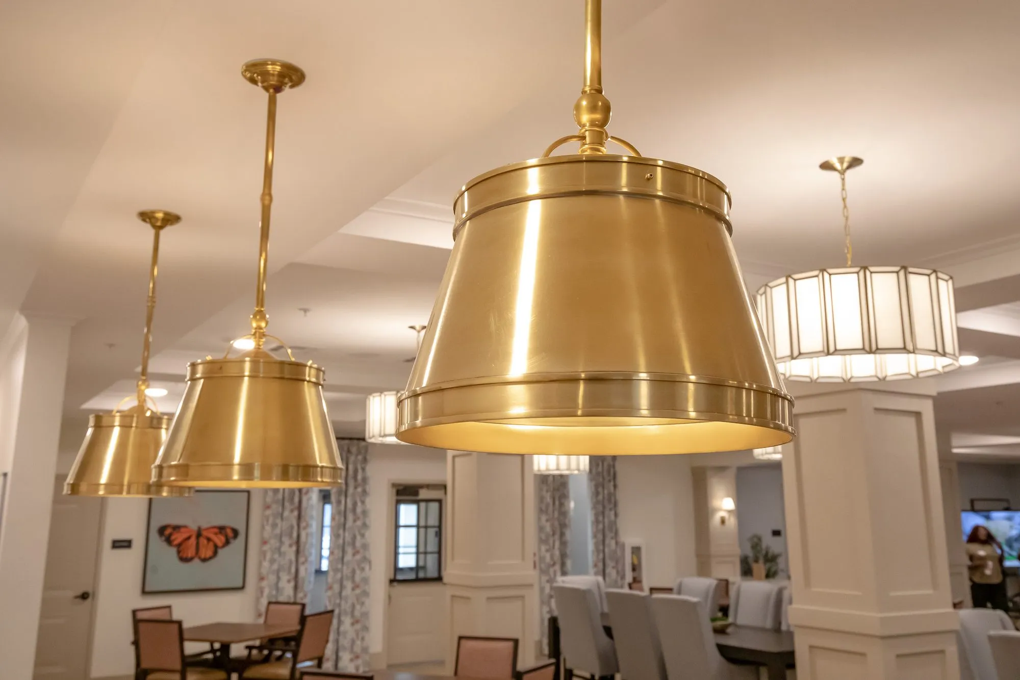 Senior living dining room with brass drum pendants hanging in a row, a butterfly painting and columns beyond