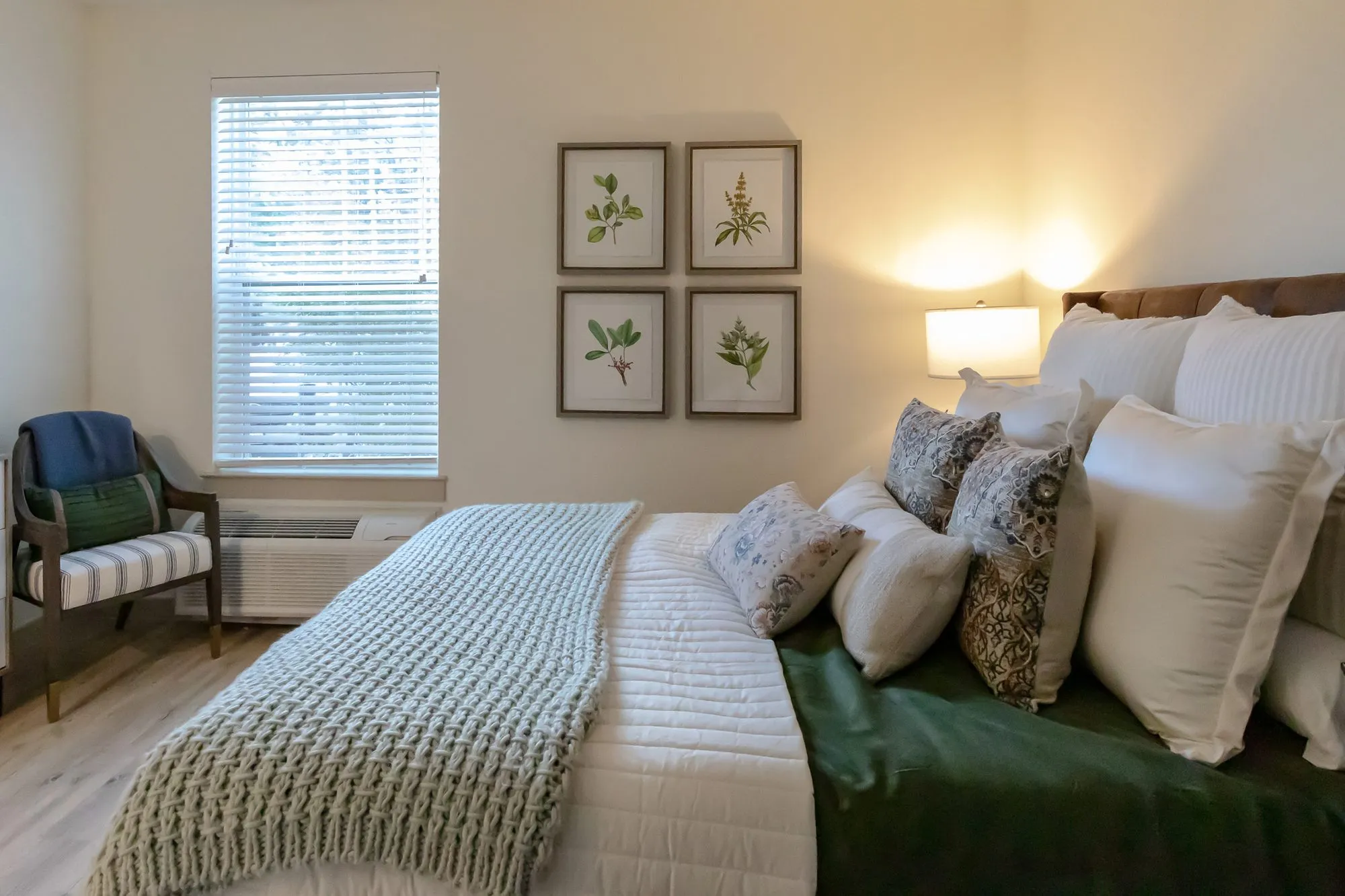 Senior living model bedroom with white bedding, a green throw and knit blanket, botanical prints and a striped chair