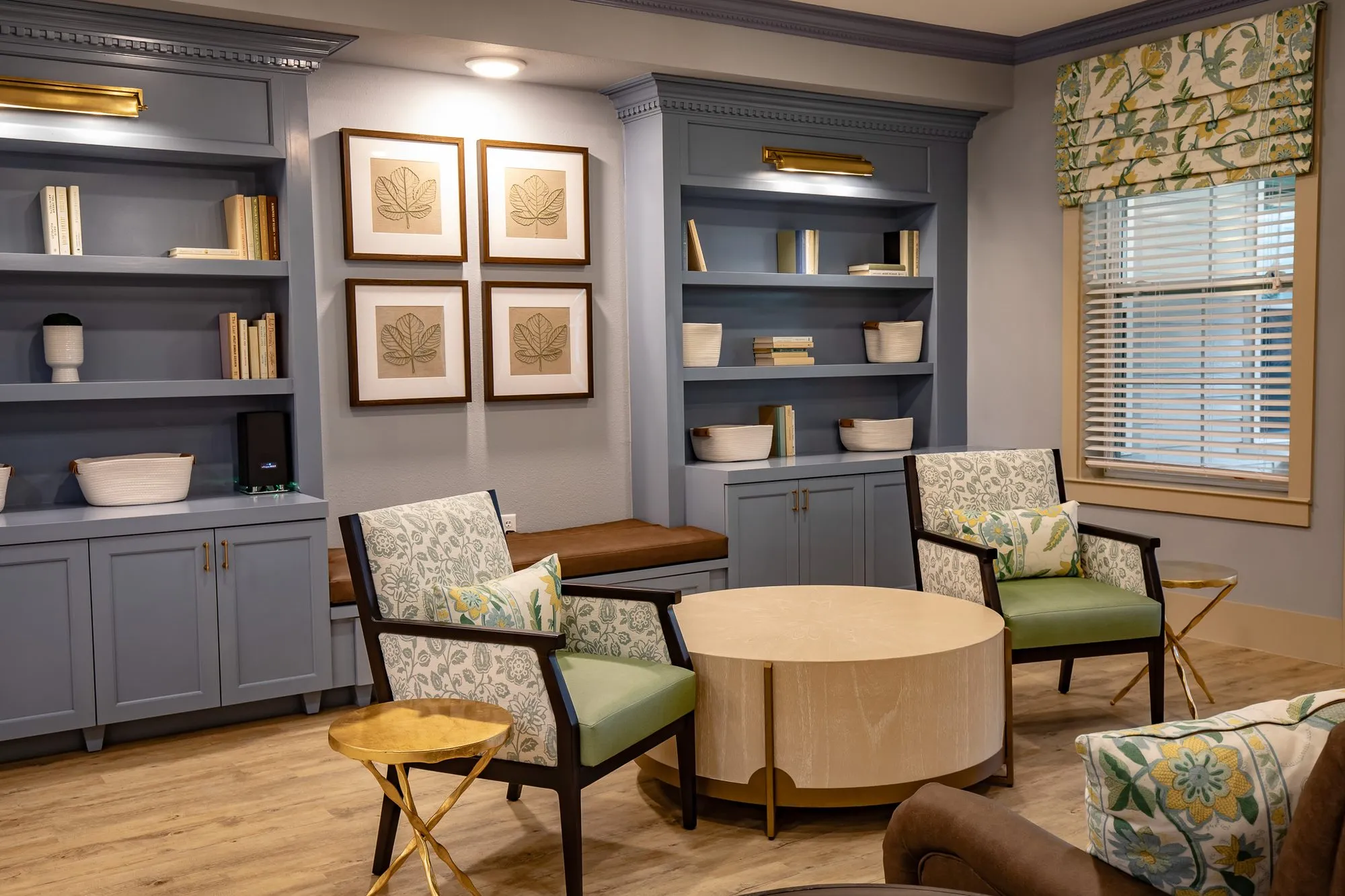 Senior living library corner with blue grey built-ins, four leaf prints and patterned armchairs around a pale wood table